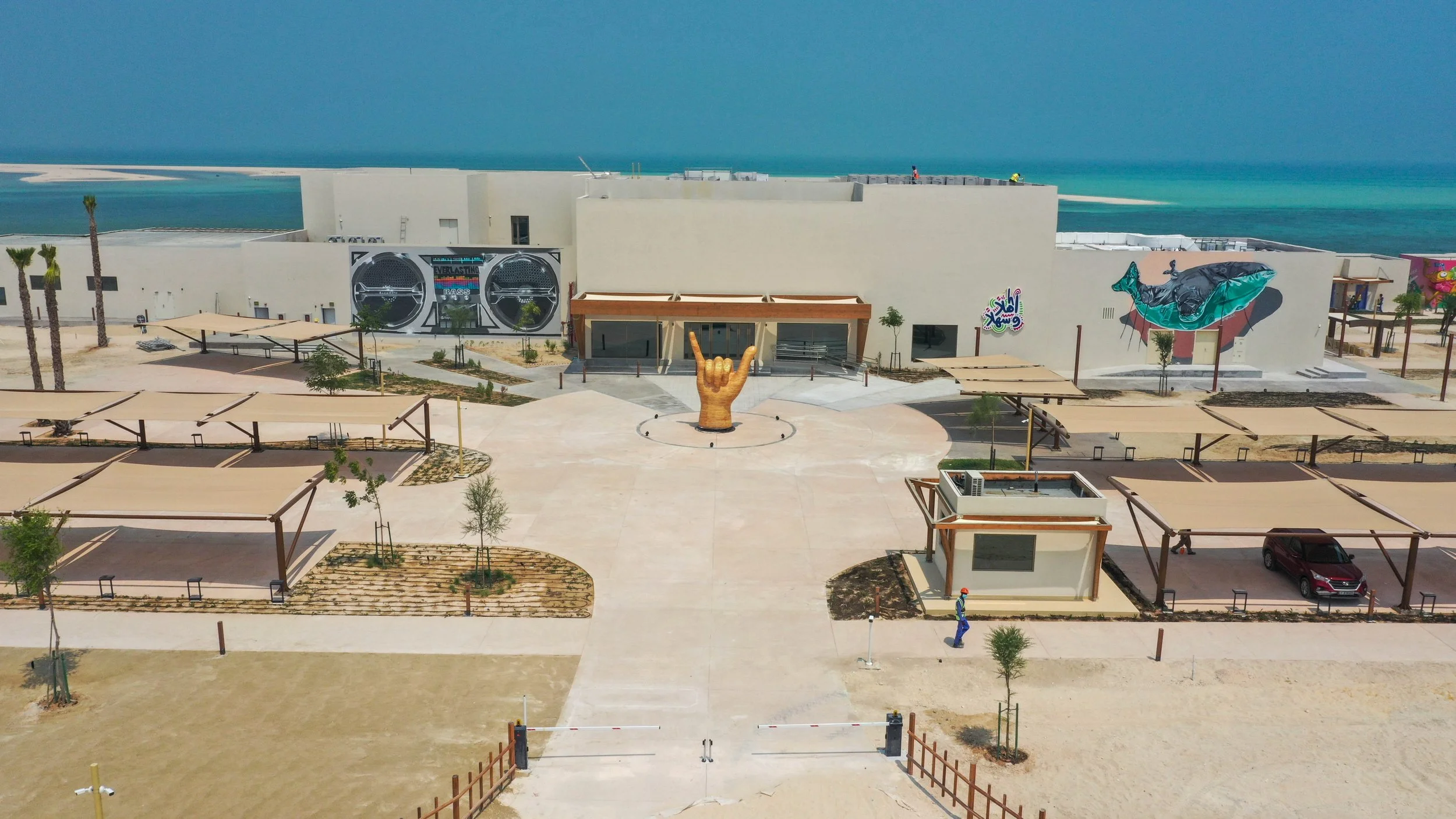 Aerial view of a white building with murals, a large golden hand sculpture, partly shaded parking area, and a sandy beach and ocean in the background.