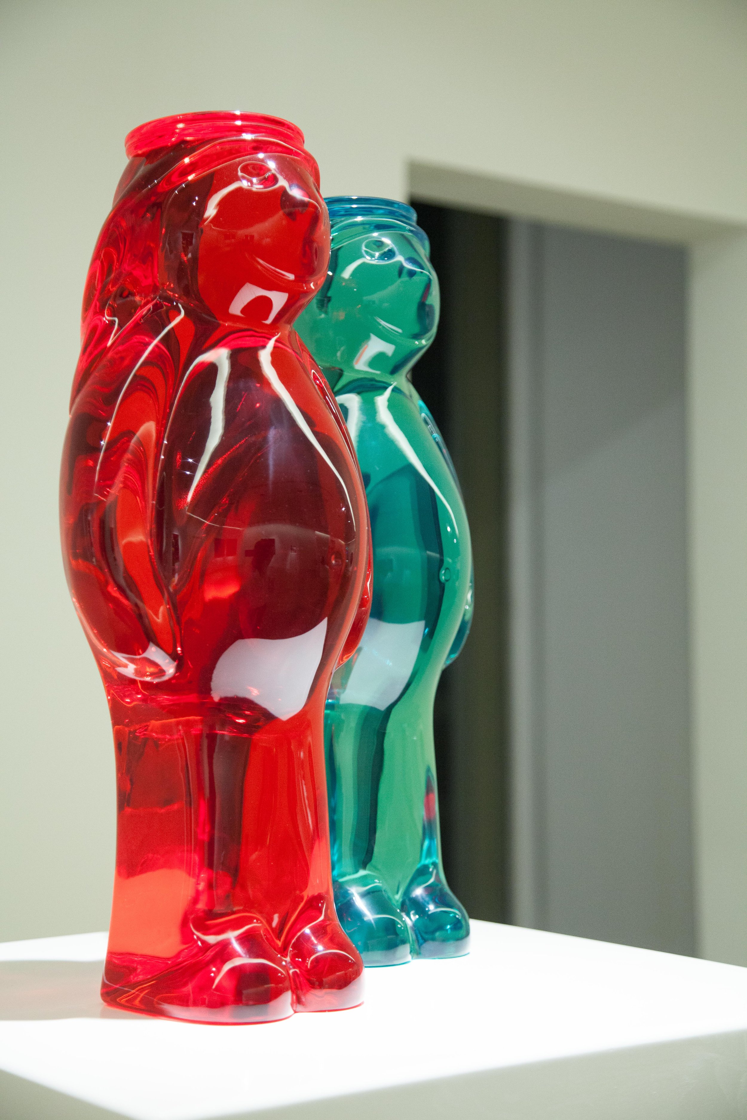 Two colorful glass teddy bear sculptures, one red and one teal, displayed on a white pedestal in an indoor setting.