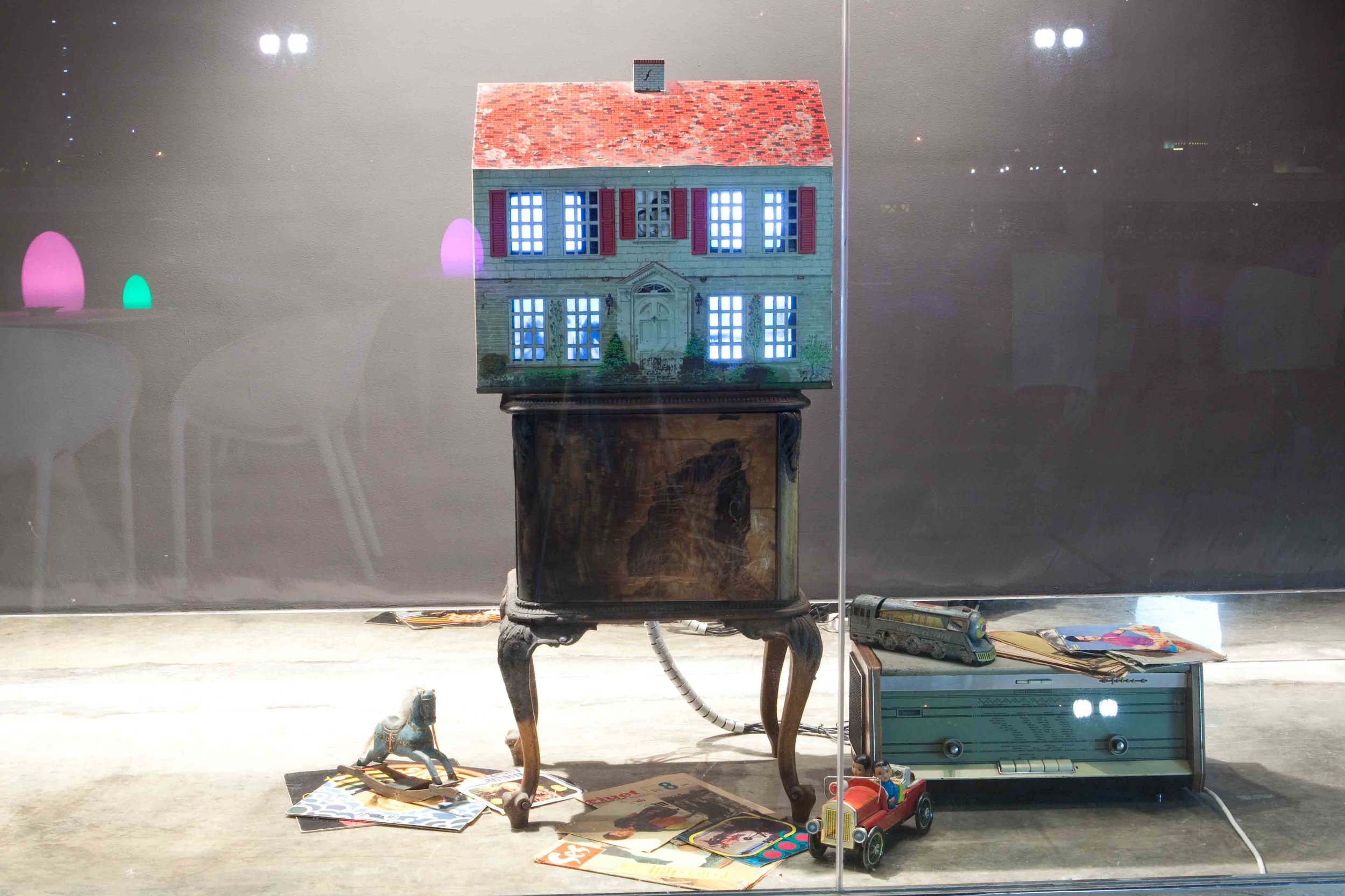 Display of a model house with multiple lit windows on an antique wooden table. Surrounding items include a vintage radio, toy horse, magazines, a toy car with two dolls, and a model train on top of the radio, all placed on the floor in front of the w