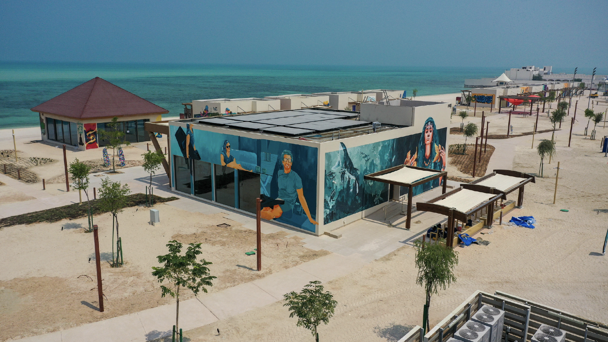 Aerial view of a beachside area with modern buildings, murals, trees, and walkways near the ocean.