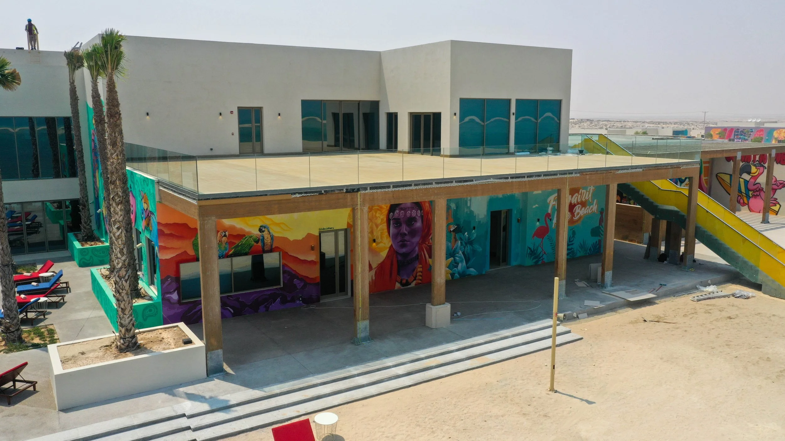 A modern building with colorful murals on the exterior walls, including images of people, flamingos, and abstract designs, with an outdoor staircase and sandy ground.
