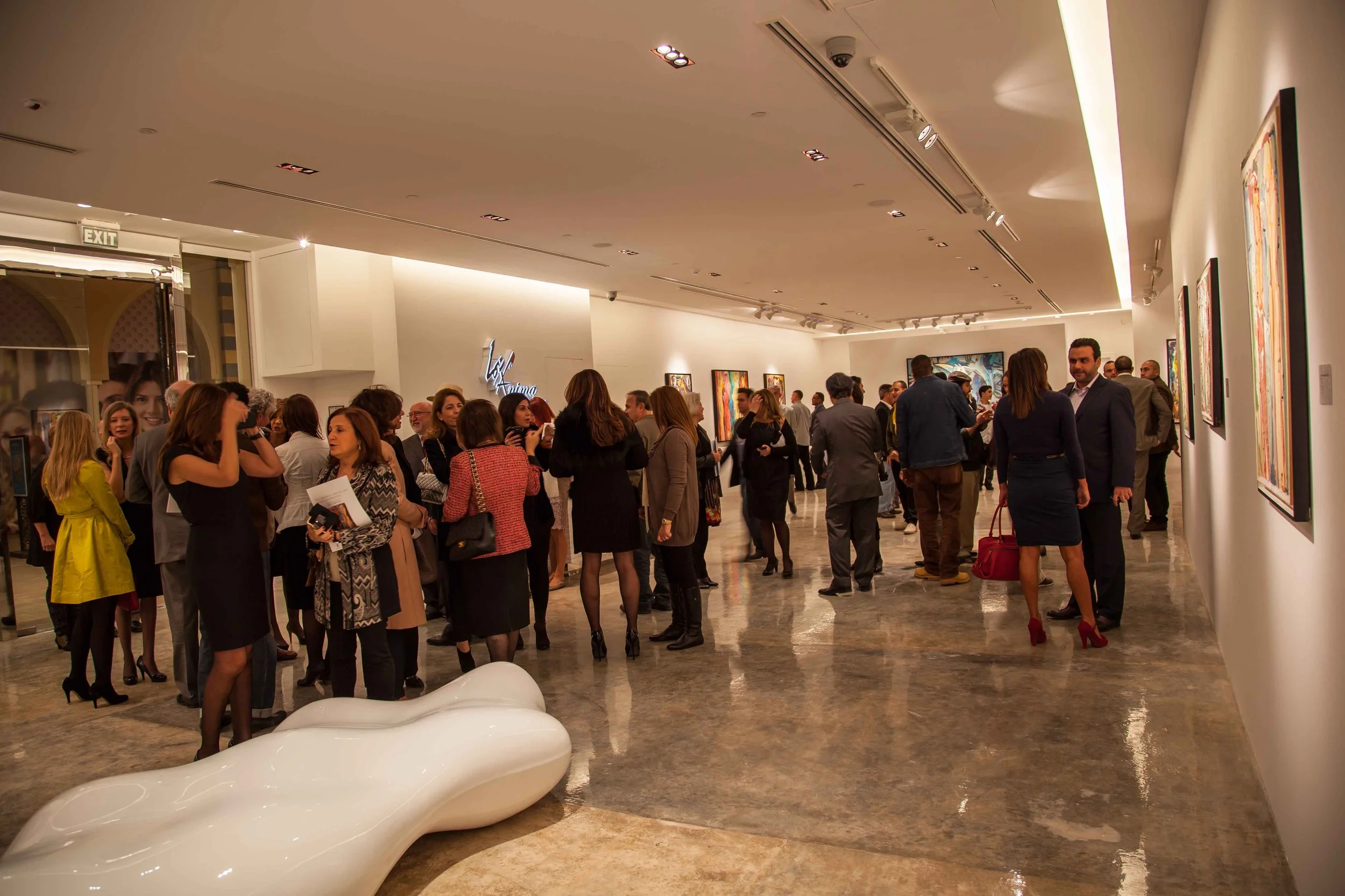 People attending an art gallery exhibition, viewing and discussing colorful paintings on white walls, with a modern white sculpture in the foreground.