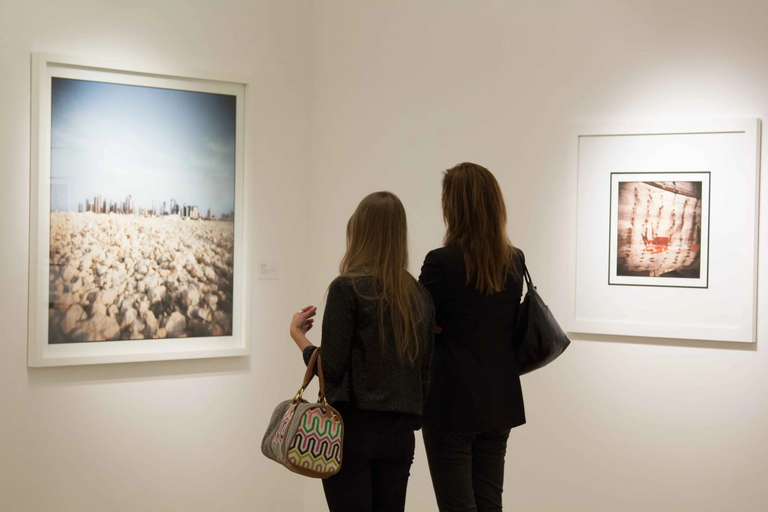 Two women in black jackets viewing artwork in a gallery, one carrying a colorful patterned bag.