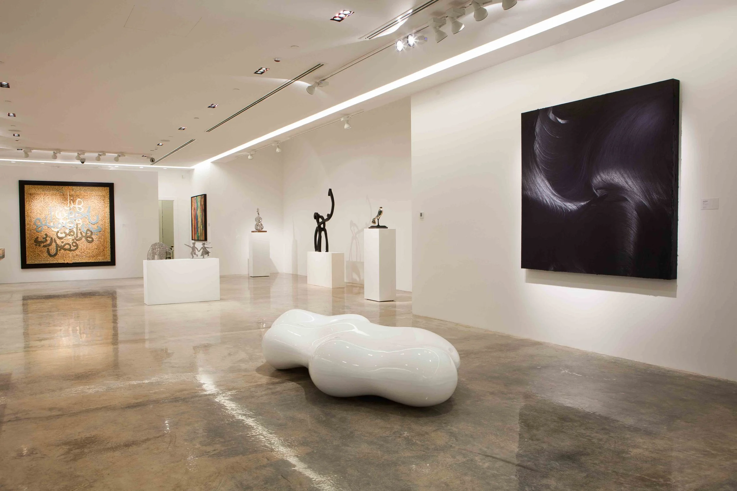 Interior of an art gallery with various sculptures and paintings. There are white walls, a polished concrete floor, and spotlights illuminating the artworks. In the foreground, a glossy white abstract sculpture resembling a bean. On the walls, large 
