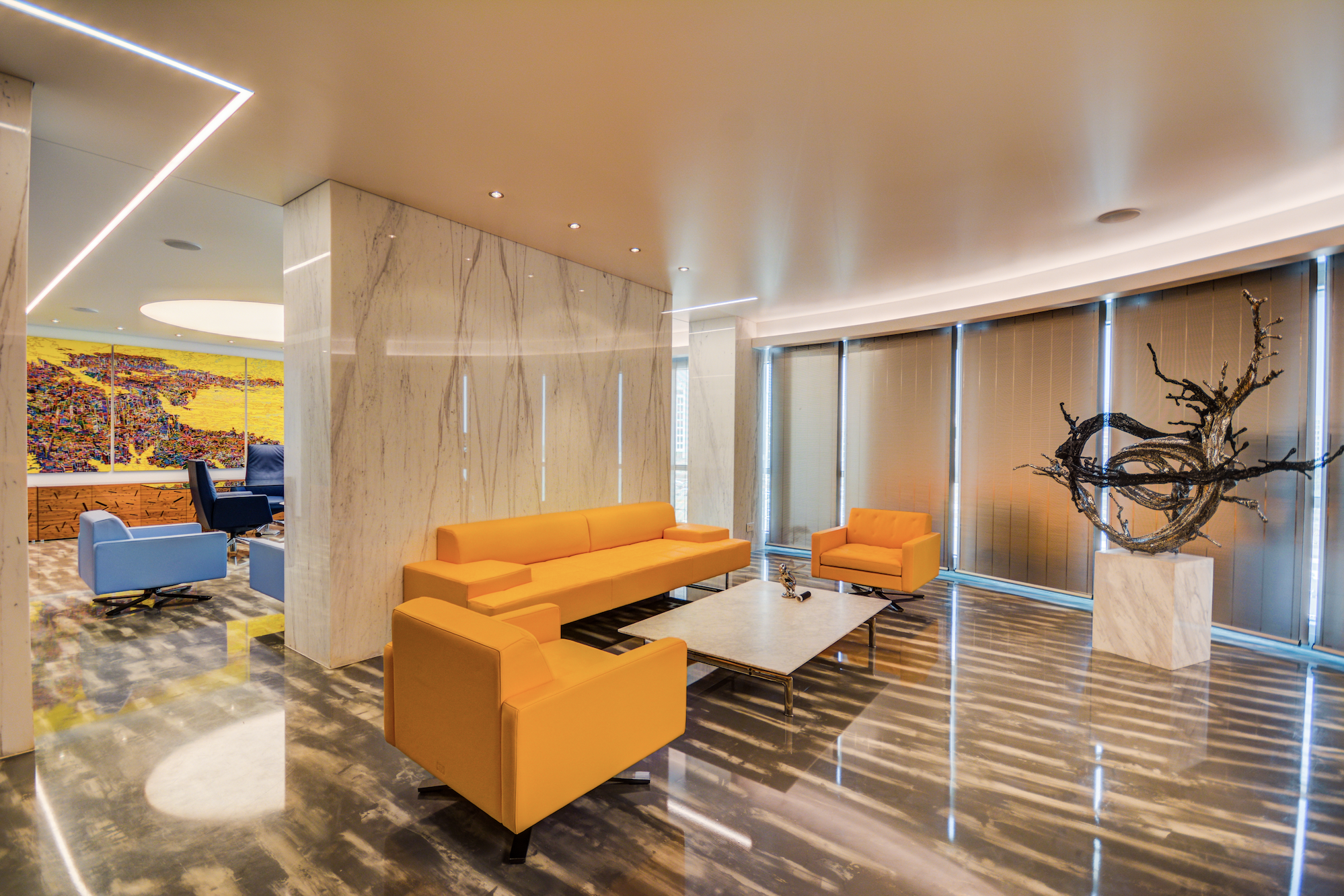 Modern hotel lobby with yellow sofas, marble floors, a contemporary sculpture, and large windows with vertical blinds.