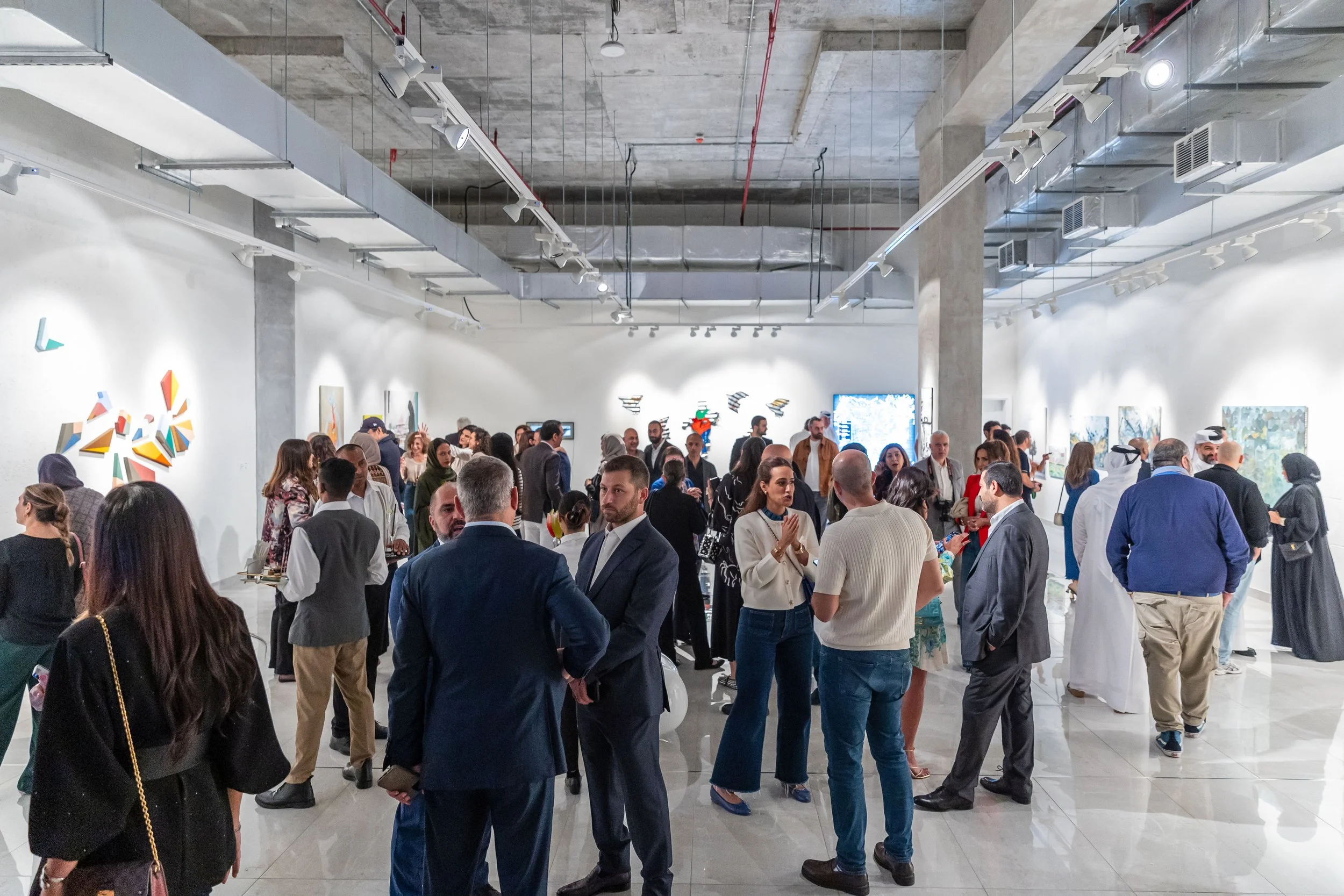 People gather at an art gallery or exhibition space, viewing and discussing artworks displayed on white walls.