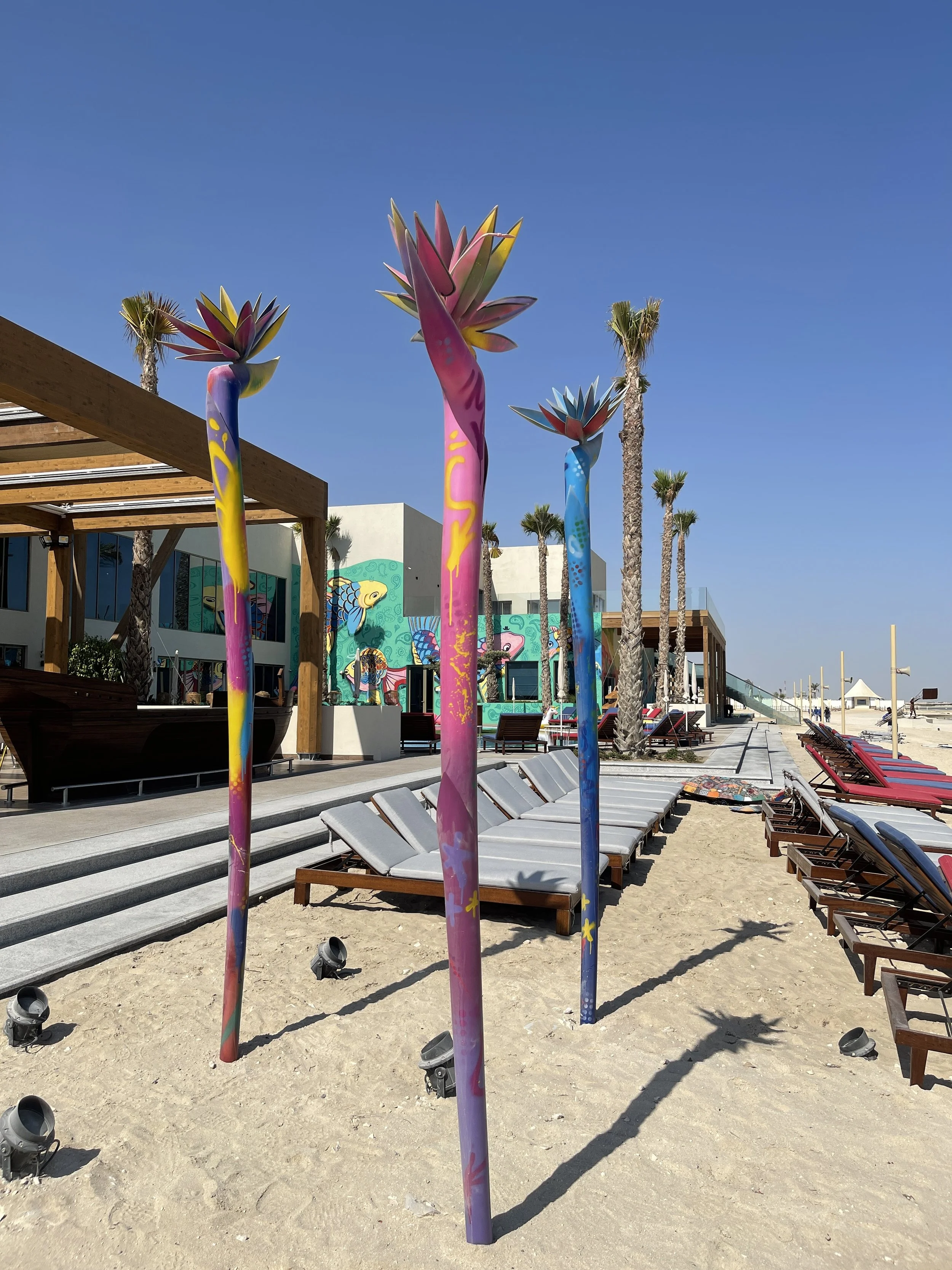 Colorful sculpture of tall, slender stems with lotus-shaped flowers at the top, on a sandy beach with lounge chairs, palm trees, and a mural-covered building in the background.
