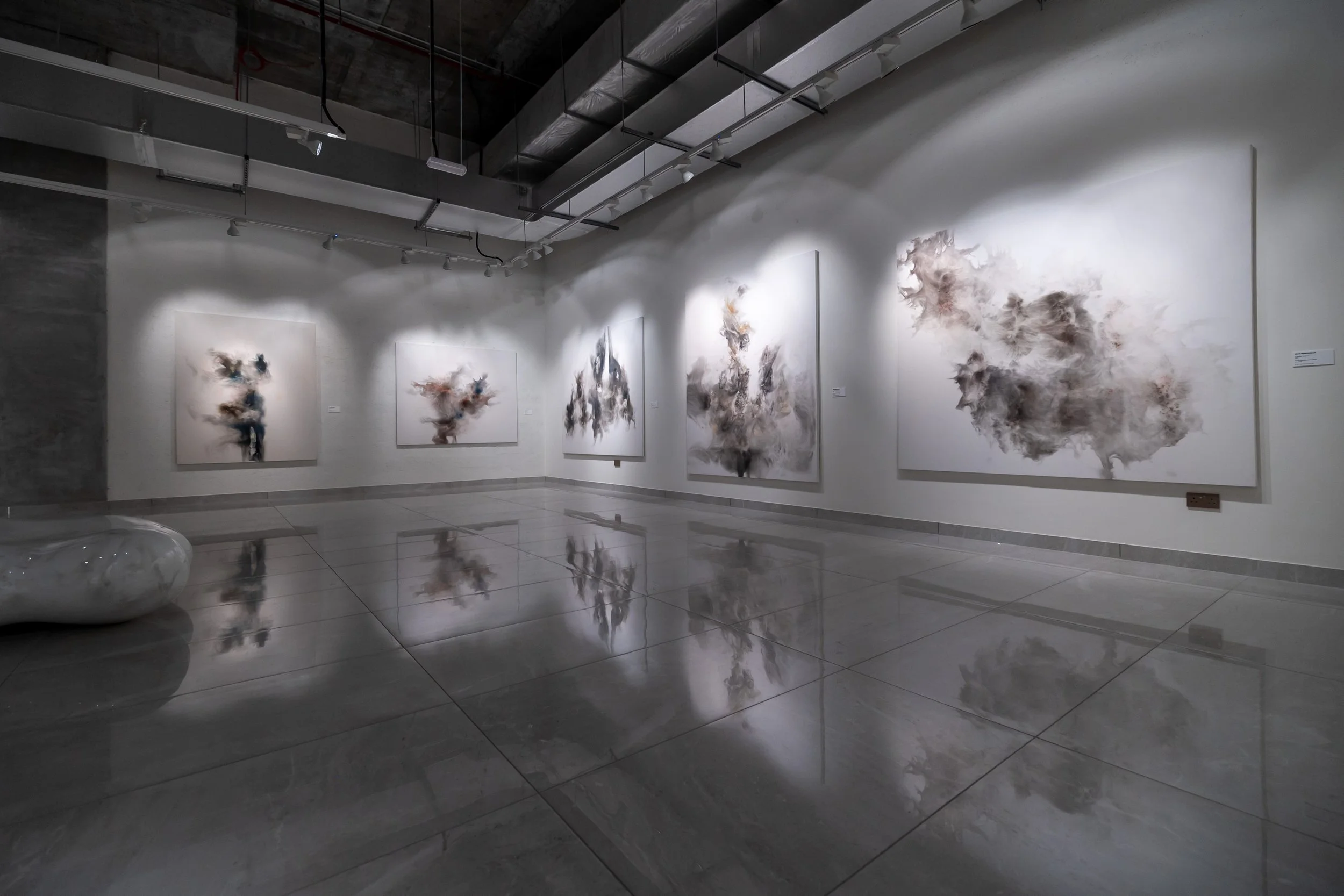 An art gallery with five abstract paintings on a white wall, illuminated by spotlights. The paintings feature cloud-like, smoky textures in neutral tones. The gallery has a polished reflective floor and a minimalistic design.