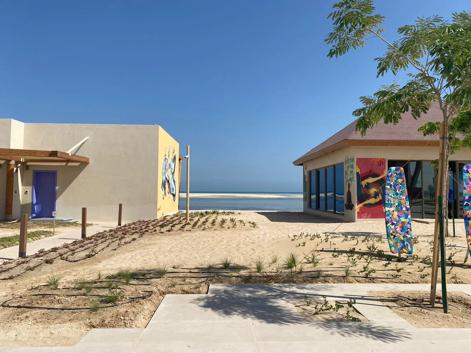 A beachside scene with two modern buildings, painted murals on walls, a sandy pathway, colorful surfboards, and ocean in the background under a clear blue sky.