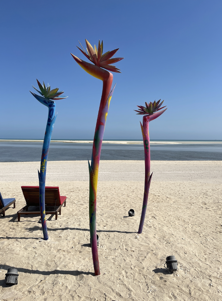 Colorful metal sculptures of tall succulent plants on a sandy beach with lounge chairs, ocean, and blue sky in the background.