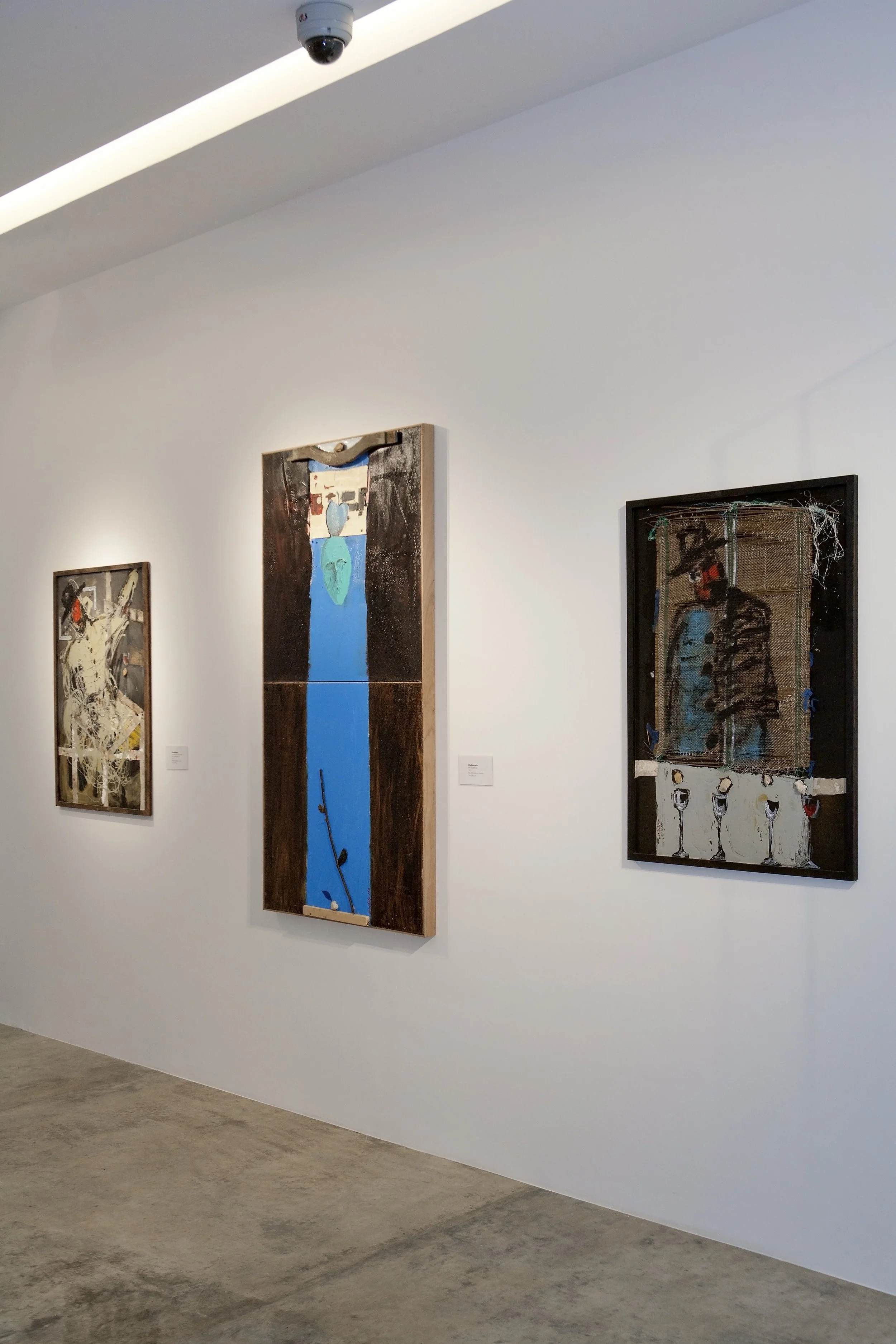 Three contemporary art paintings hanging on a white gallery wall, with a security camera mounted on the ceiling.