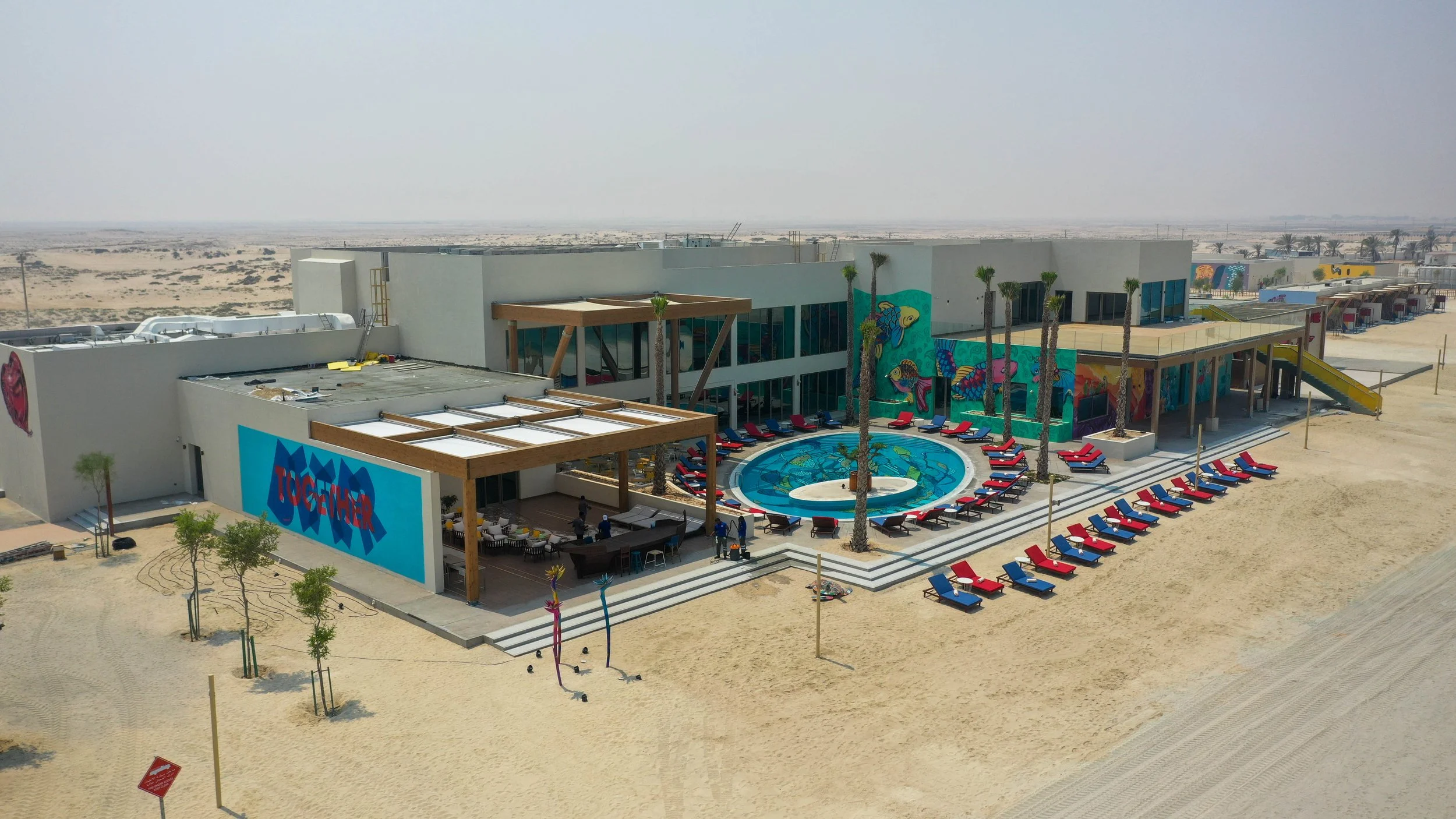 Aerial view of a modern beachfront building with a swimming pool, surrounded by red and blue lounge chairs, palm trees, and colorful wall murals, in a desert landscape.