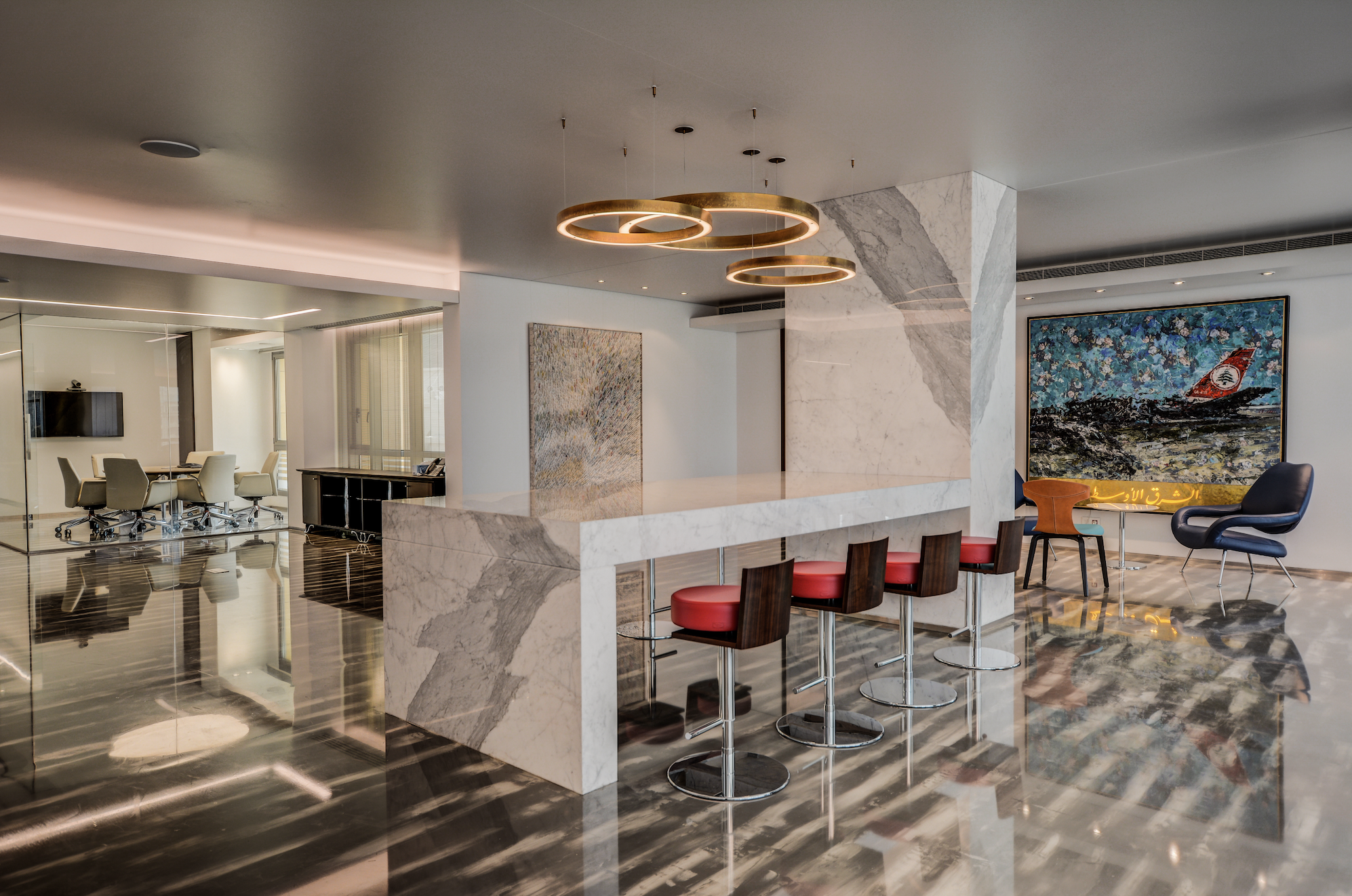 Modern office lobby featuring a marble bar with four red stools, abstract artworks on the walls, a seating area with a large painting, and a conference room with an oval table and cream chairs.