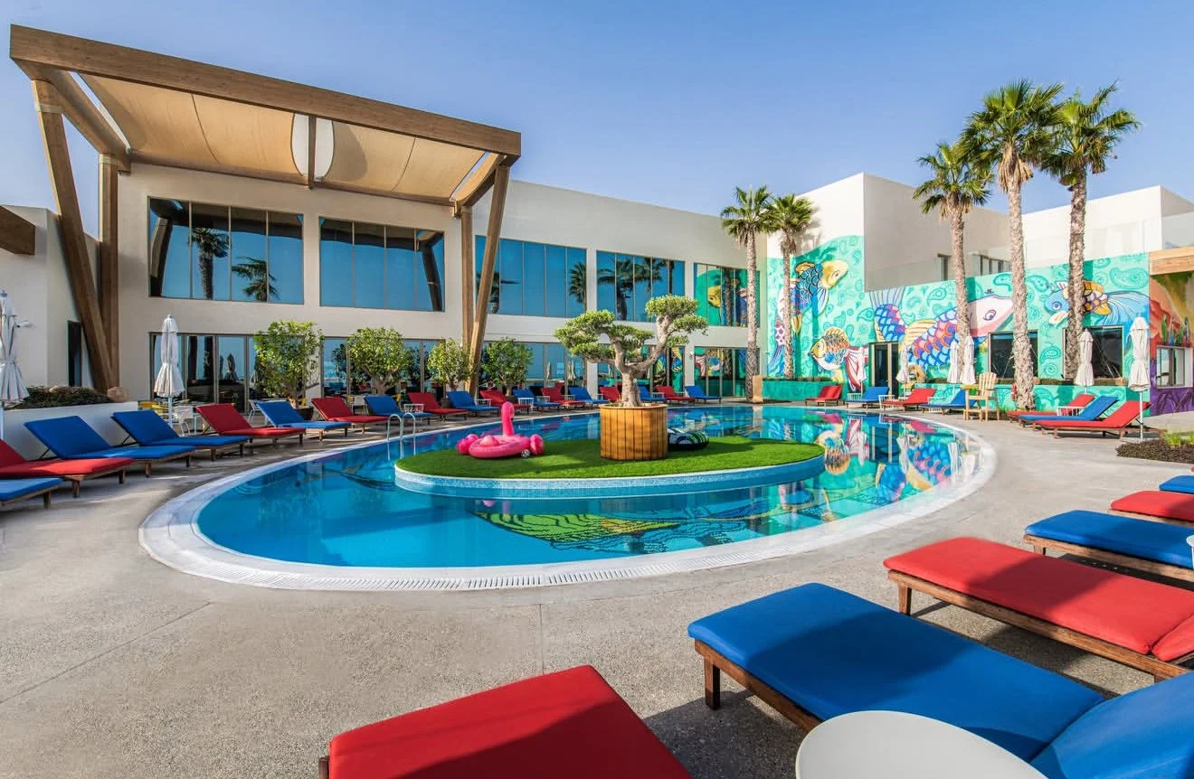 Empty outdoor swimming pool surrounded by red and blue lounge chairs, with a small island in the pool featuring a bonsai tree, pink flamingo float, and a pair of black sunglasses. There are tall palm trees and colorful fish and ocean-themed murals on