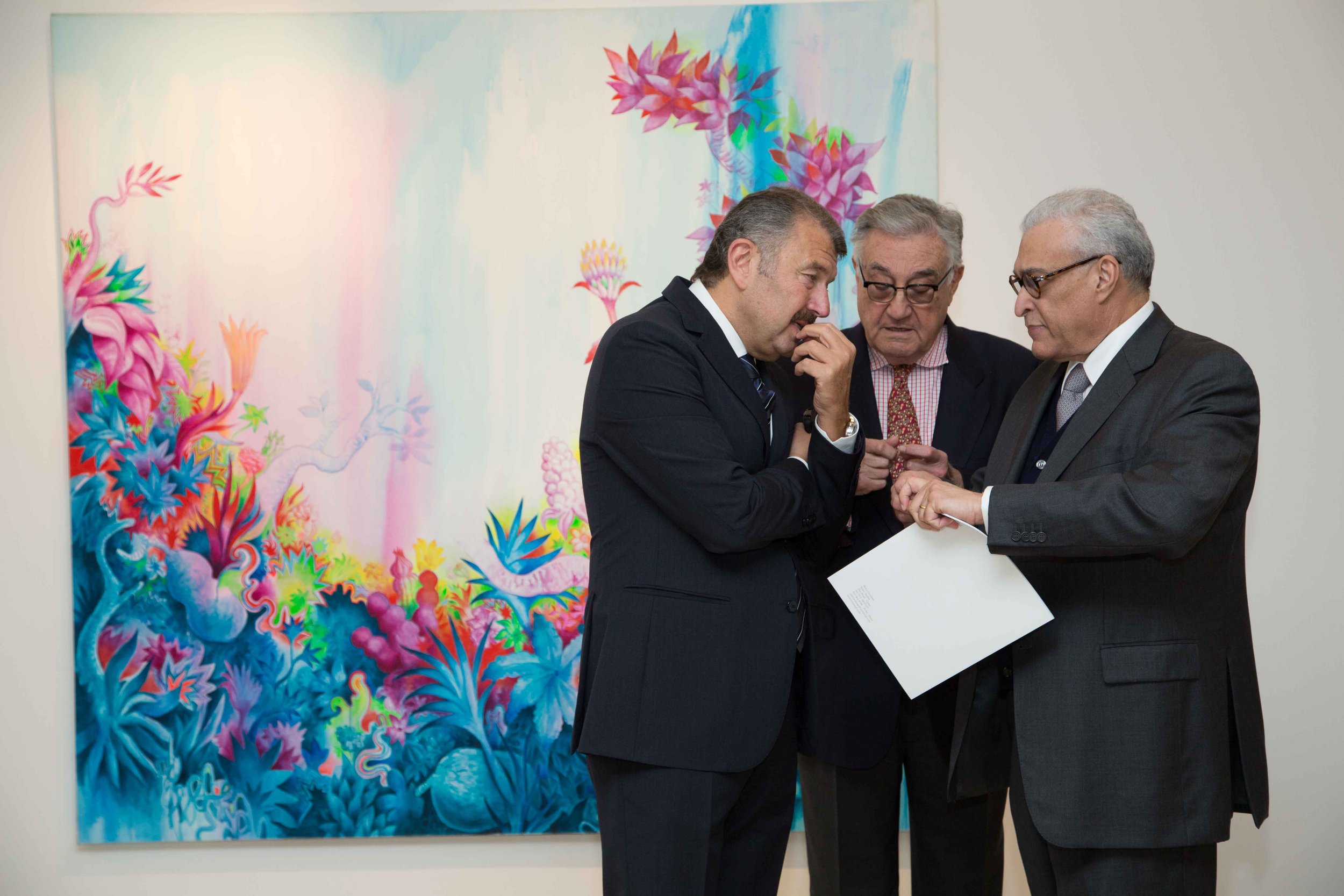 Three men in business suits stand and look at a document in front of a colorful abstract artwork in a gallery.