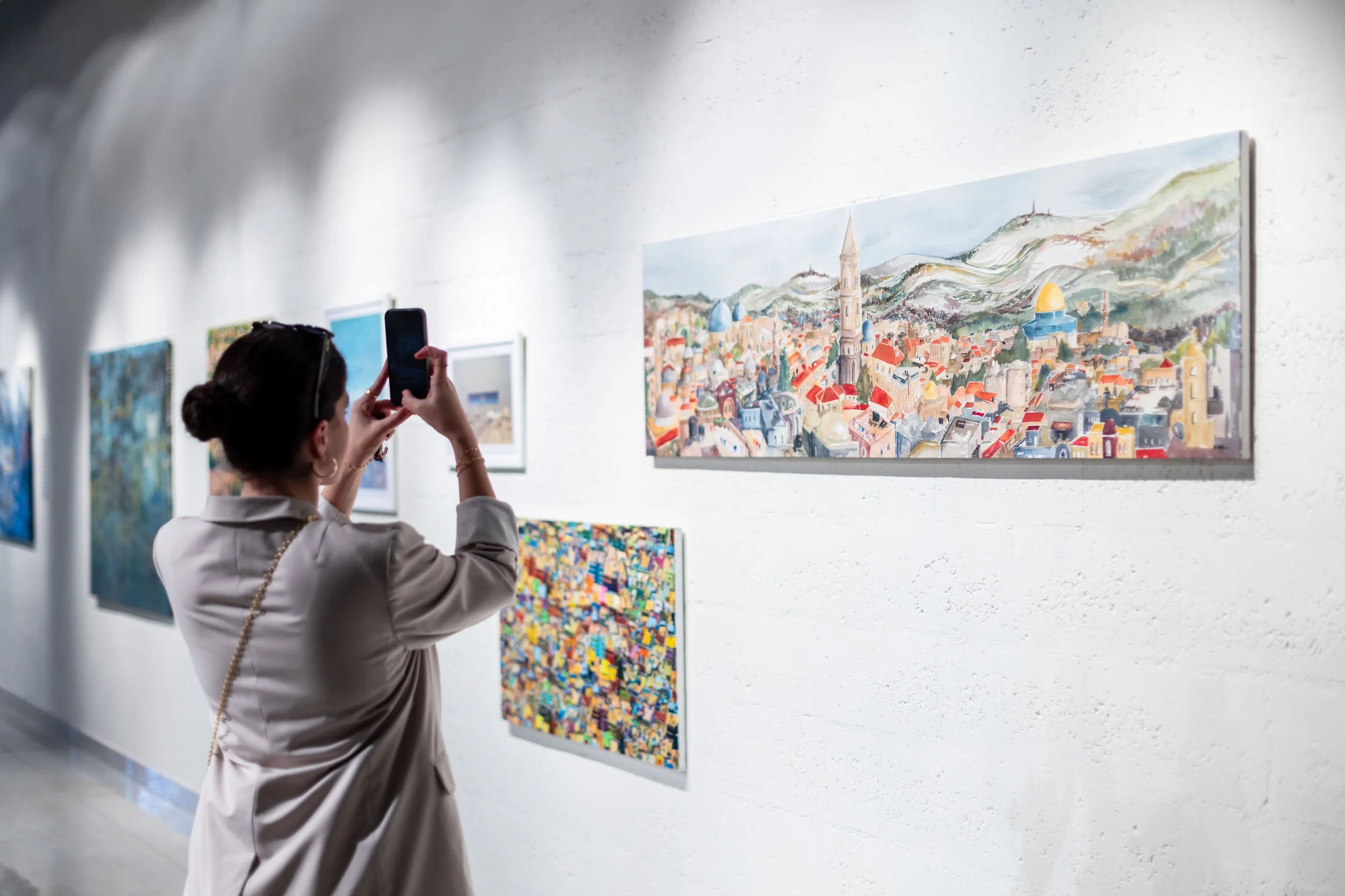 A woman with dark hair in a bun, wearing a beige jacket, is taking a photo of a painting with her smartphone inside an art gallery. The wall displays multiple artworks, with a colorful cityscape painting featuring a tall clock tower and mountains in 