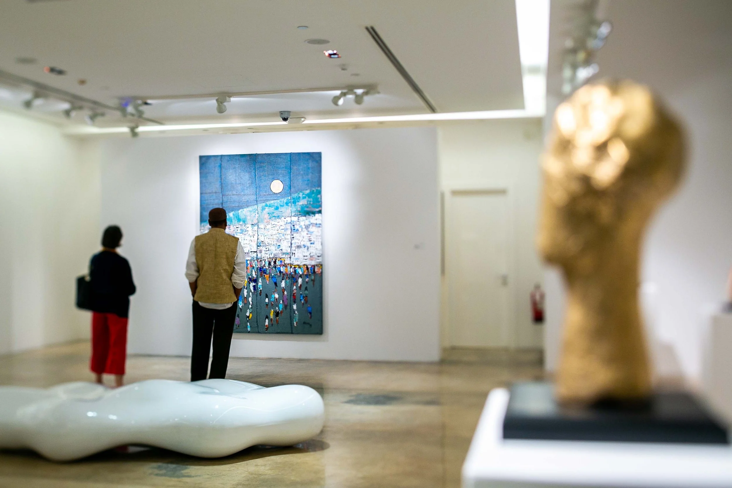 People viewing artwork in an art gallery, with a painting of a cityscape and moon on the wall, a white abstract sculpture on the floor, and a blurred gold sculpture in the foreground.