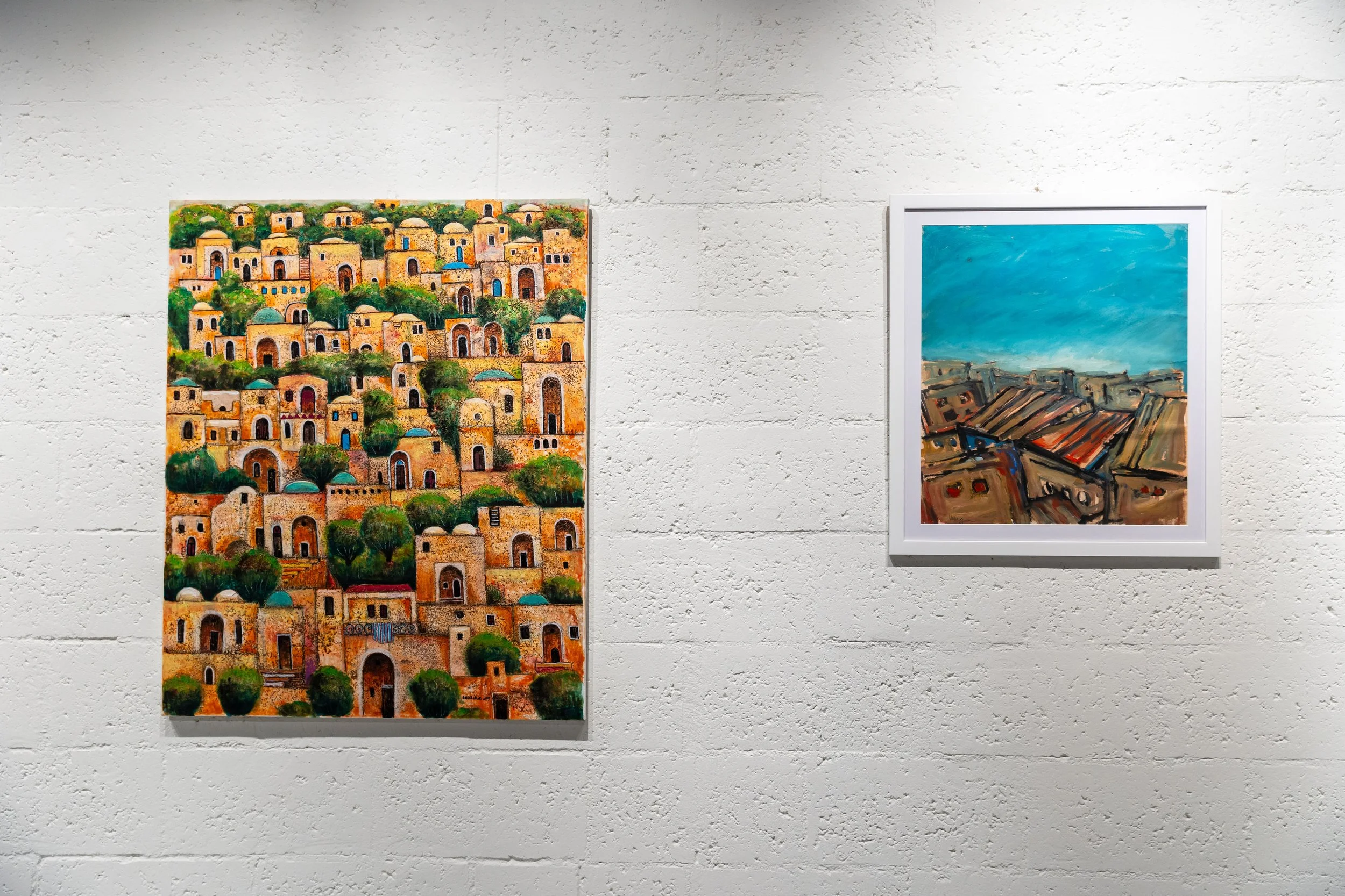 Two paintings displayed on a white brick wall, one depicting a vibrant cityscape with many small buildings and trees, and the other showing a landscape with buildings under a blue sky.