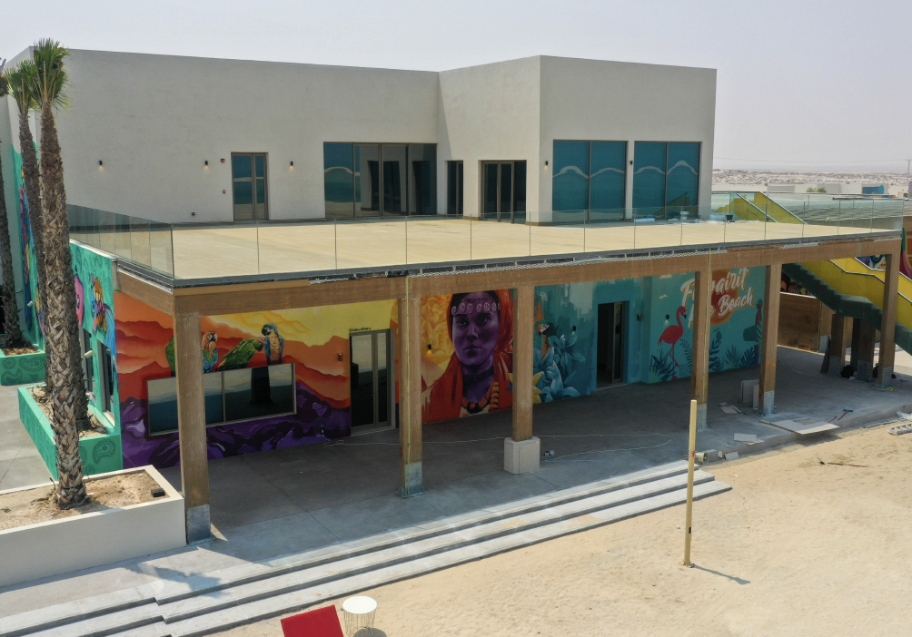 A modern two-story building with colorful murals and large glass windows, featuring a rooftop deck with a glass railing, set in a sandy area with palm trees.