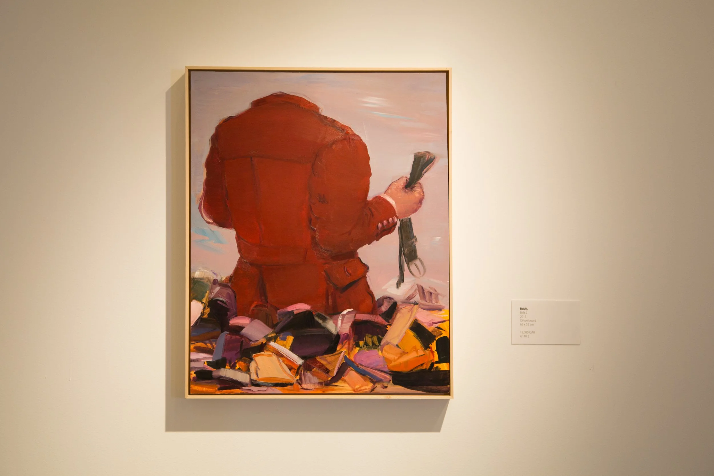 Painting of a person in a red jacket from behind, holding a large black object, standing near a pile of colored objects, with a label indicating the art piece's title and details.