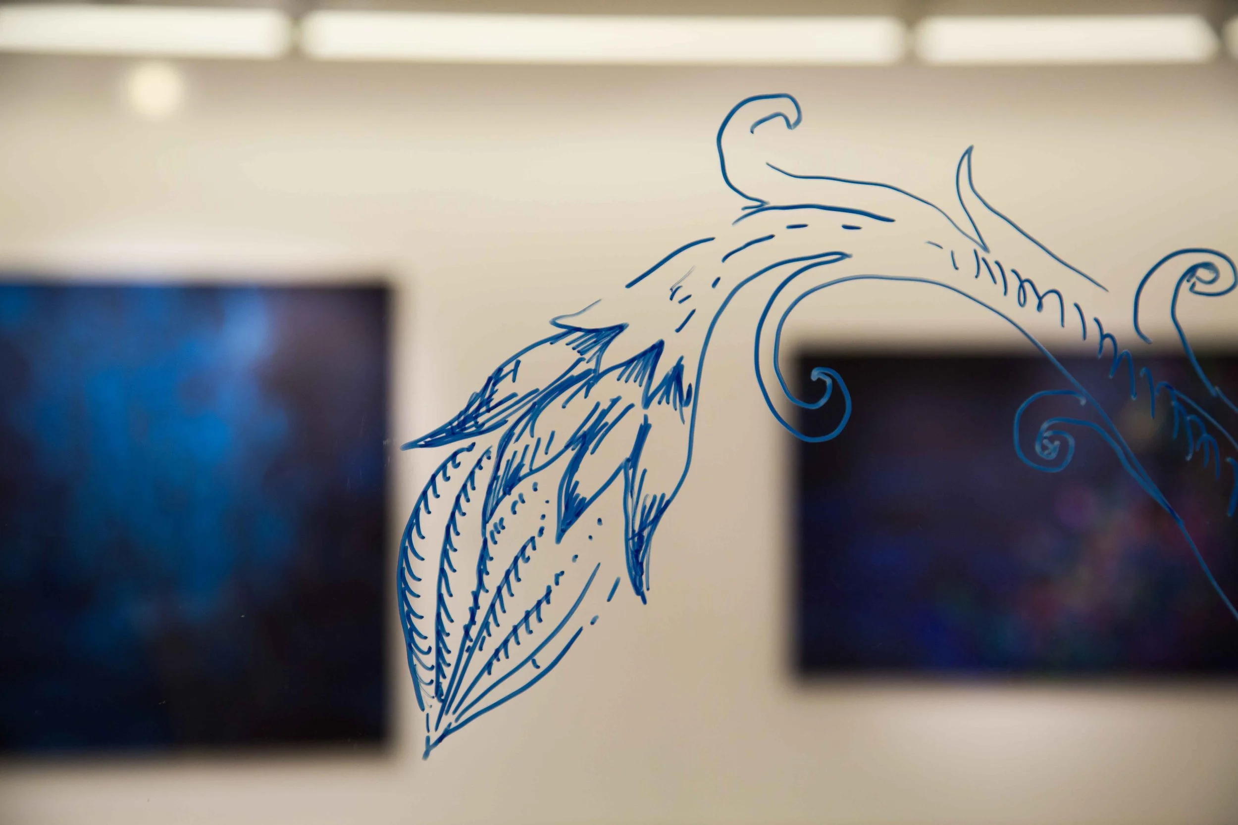 Blue line drawing of a stylized bird or dragon on a glass surface with two blurred digital screens in the background.