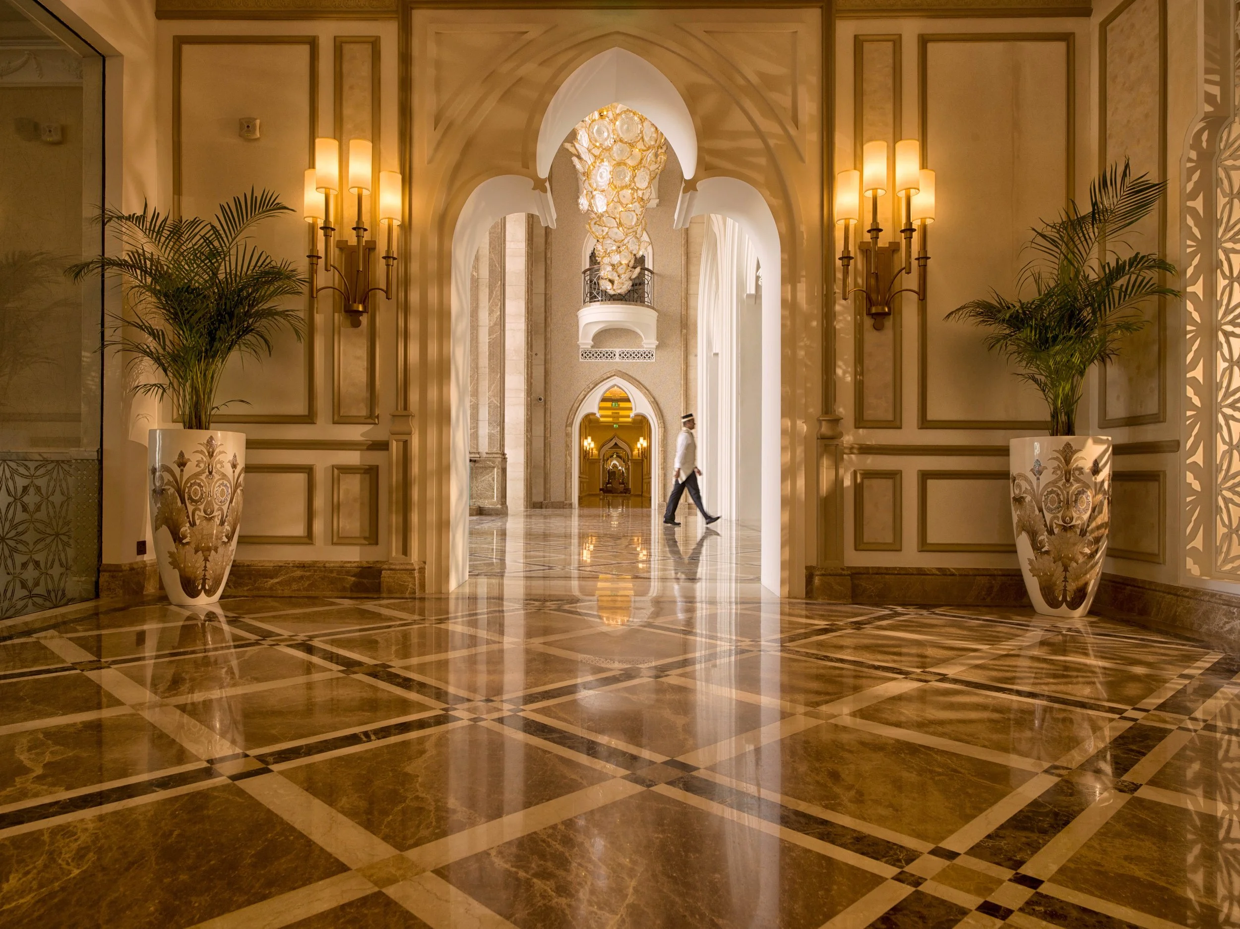 Luxurious hotel lobby with marble floors, ornate wall paneling, large potted plants, and decorative lighting fixtures, with a person walking through the archway.