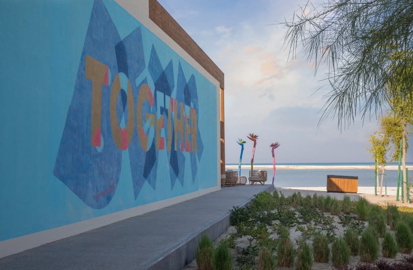 Beachside mural with the words 'TOGETHER' painted on a large wall, colorful palm trees, benches, and ornamental plants near the ocean.