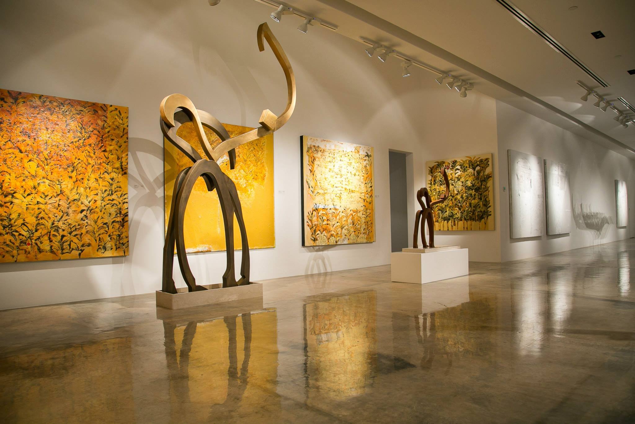 Interior of an art gallery featuring yellow paintings with abstract patterns and two modern wooden sculptures on pedestals, with reflections on a polished concrete floor.