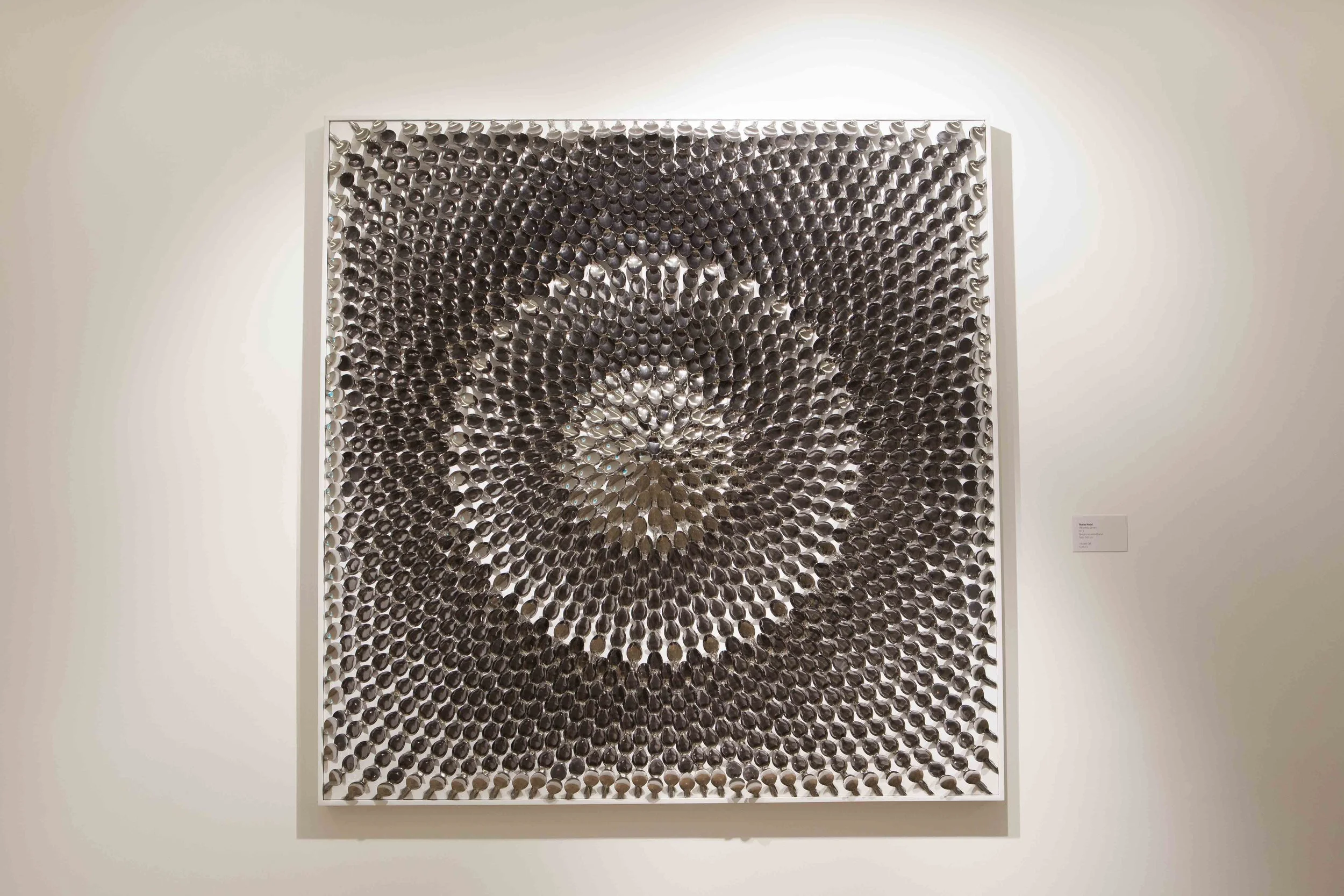 Artwork made of numerous metallic spoons arranged in a spiral pattern on a white gallery wall.