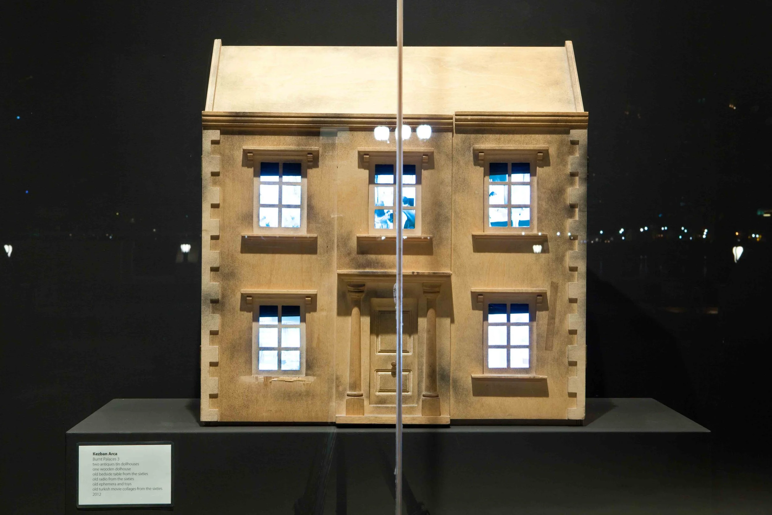 A miniature model of a two-story wooden house with six windows and a central door, displayed on a black stand in a dimly lit room.