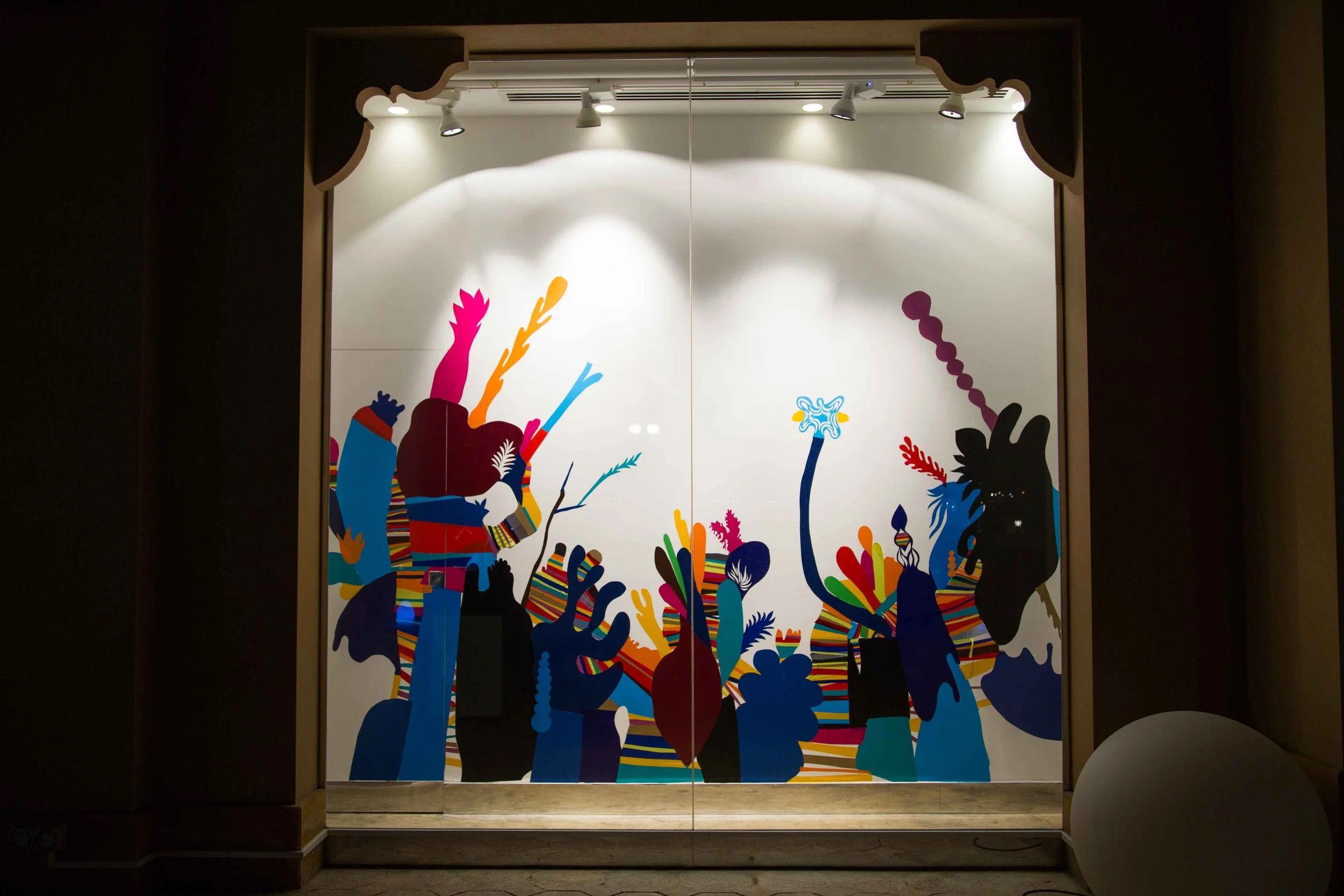 Colorful pattern of abstract shapes resembling a crowd of hands and faces on a white wall, with spotlights illuminating the display.