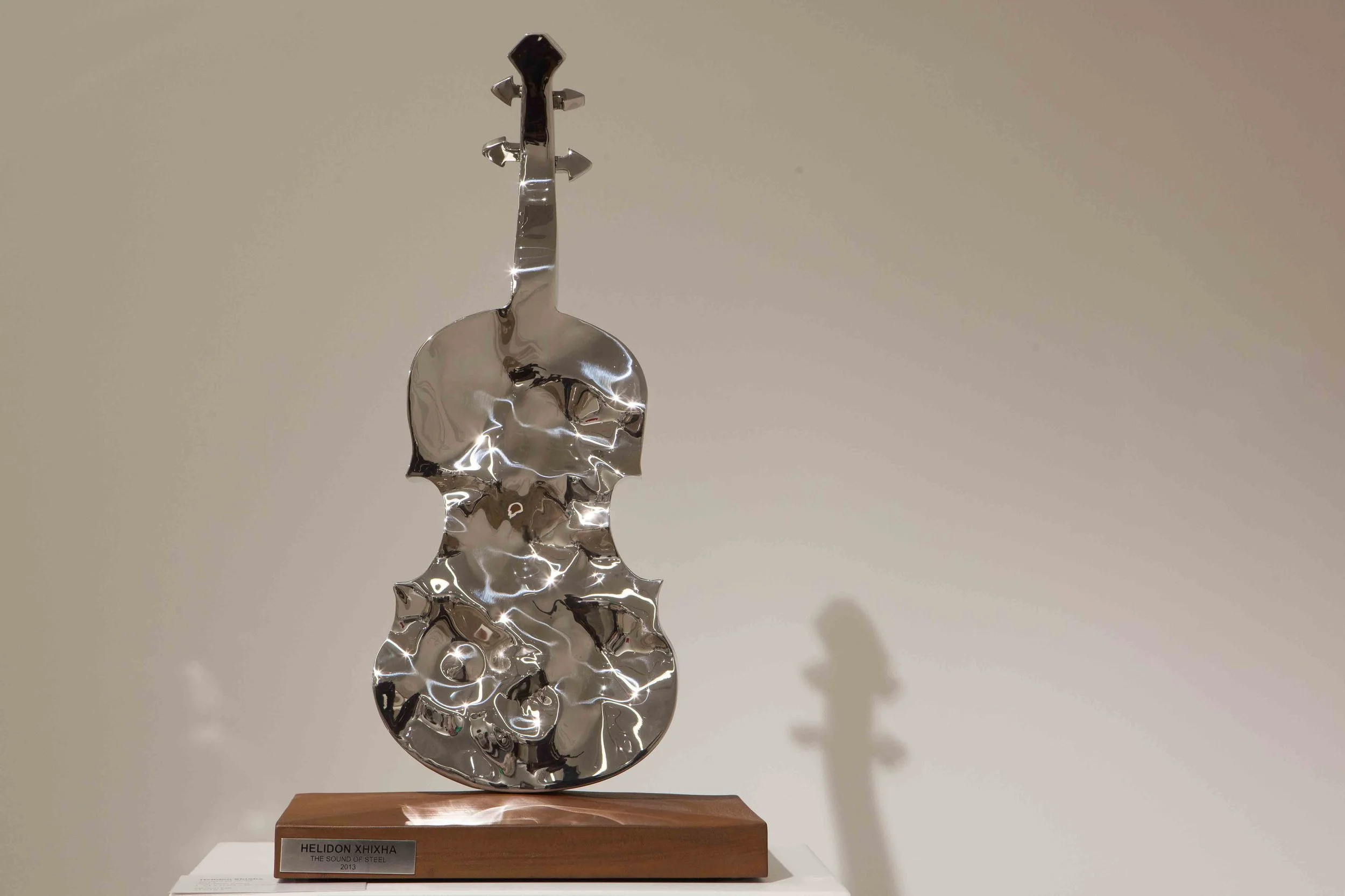 Sculpture of a stylized violin made of shiny metal, mounted on a wooden base labeled "HELIDON XHIXHA - THE SOUND OF STEEL".