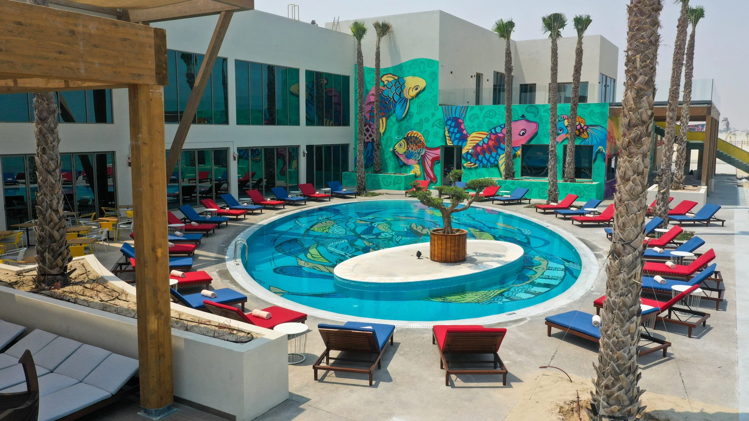 Colorful outdoor pool area with a mural of vibrant fish on the wall, surrounded by palm trees and red and blue lounge chairs.
