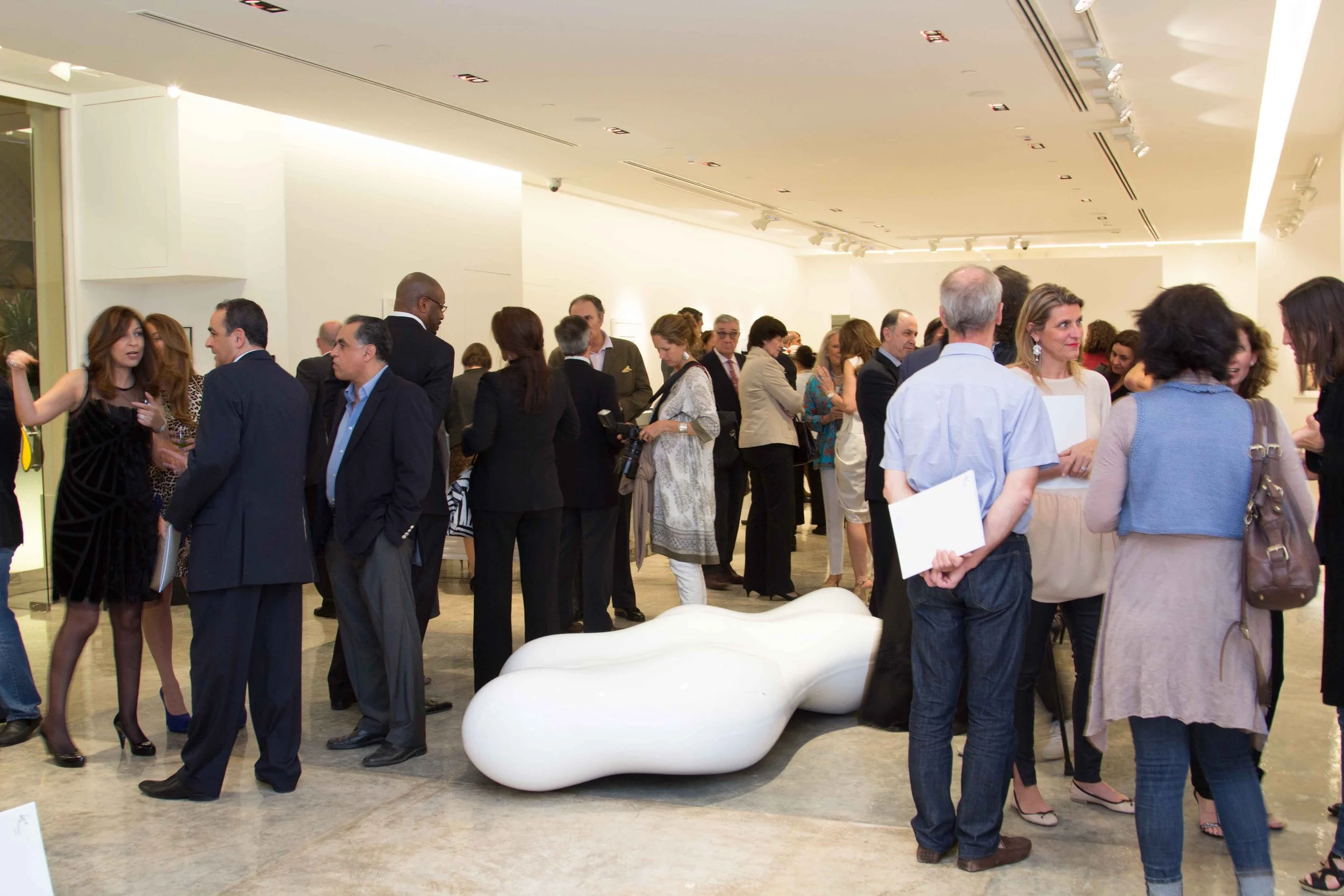 A large group of people attending a social or professional event in an art gallery or modern venue, engaged in conversations, with some holding papers or notebooks, and a white abstract sculpture on the floor.
