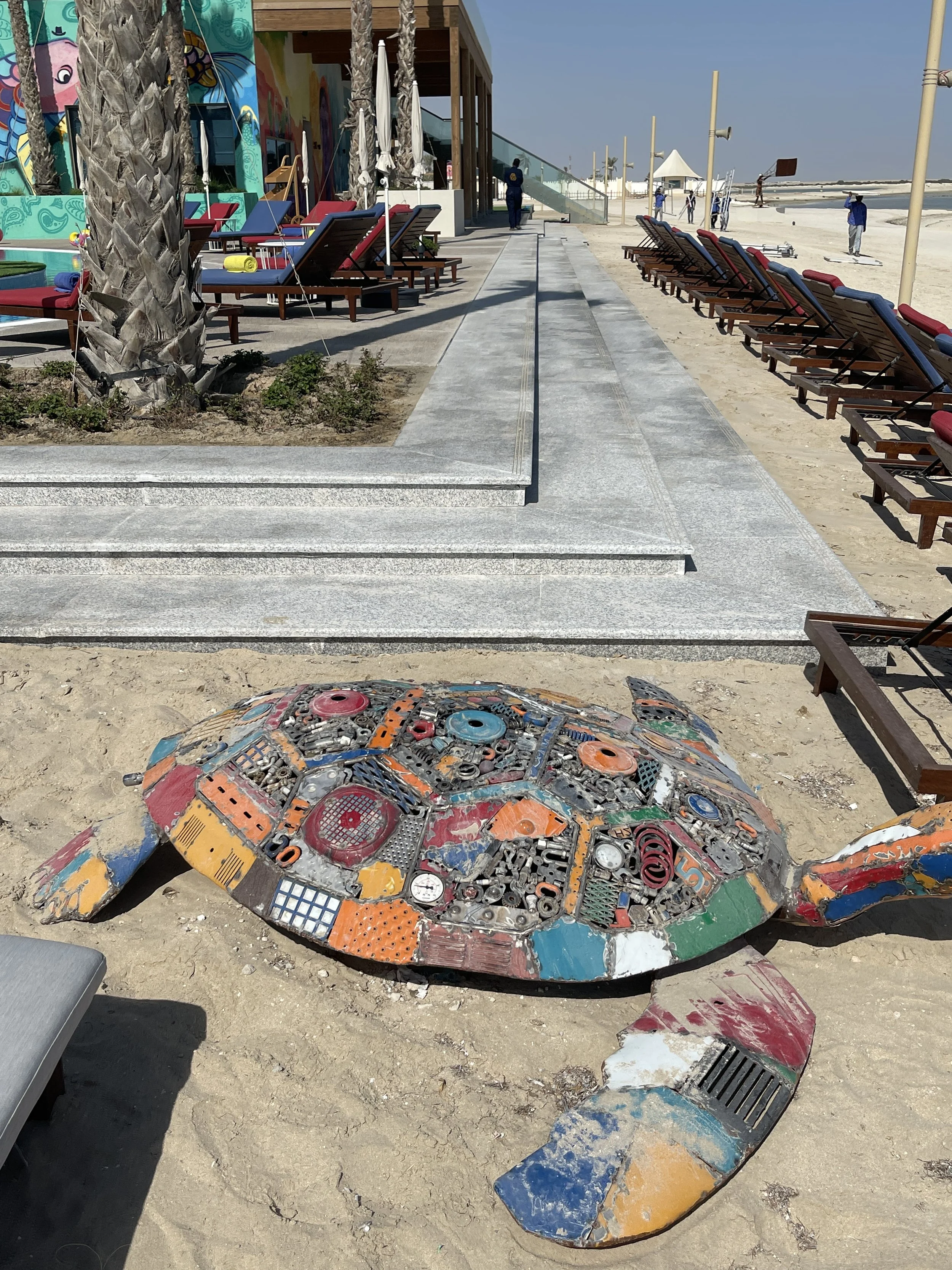 A large turtle sculpture made of colorful recycled metal parts is on the sandy beach near rows of lounge chairs, palm trees, and a building with street art.