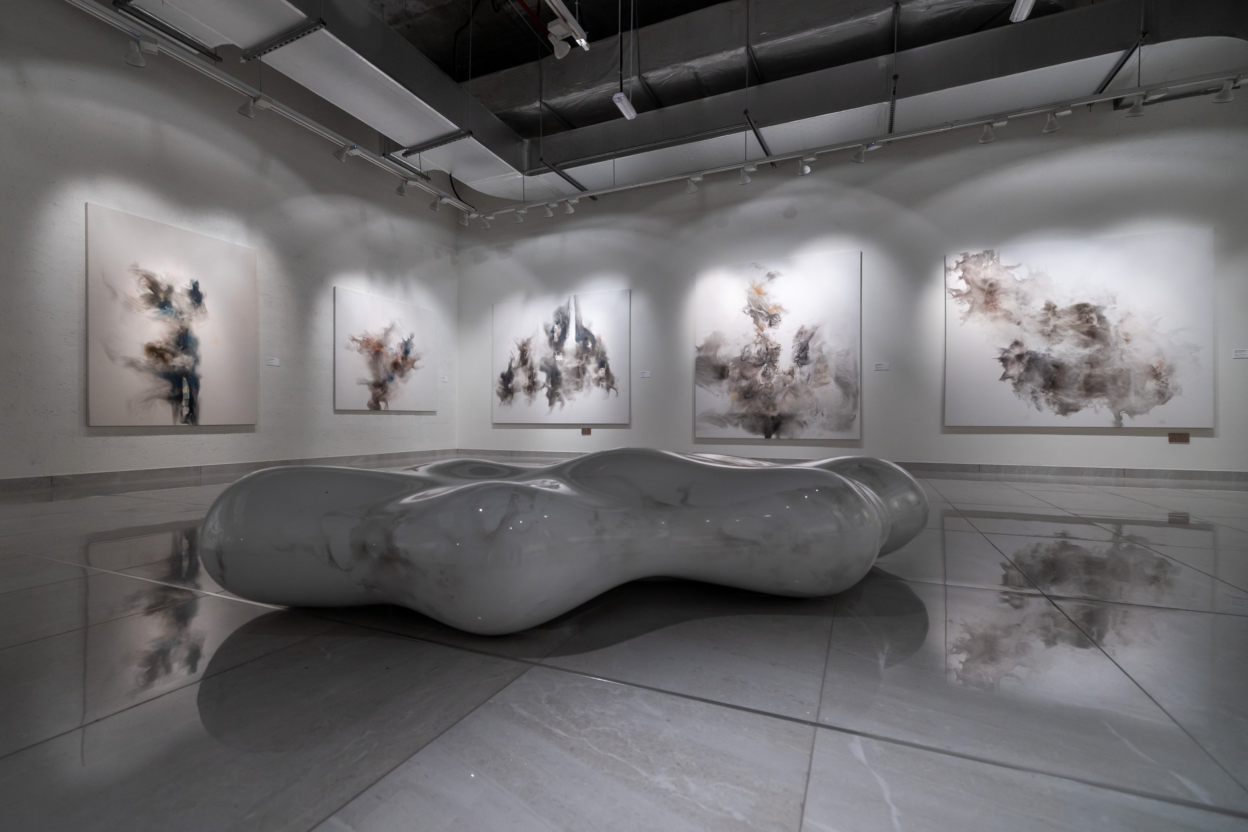 An art gallery with five abstract paintings on the white walls and a sculptural piece on the reflective floor. The paintings feature swirling, cloud-like forms in neutral tones and subtle colors. The sculpture is smooth, elongated, and irregularly sh
