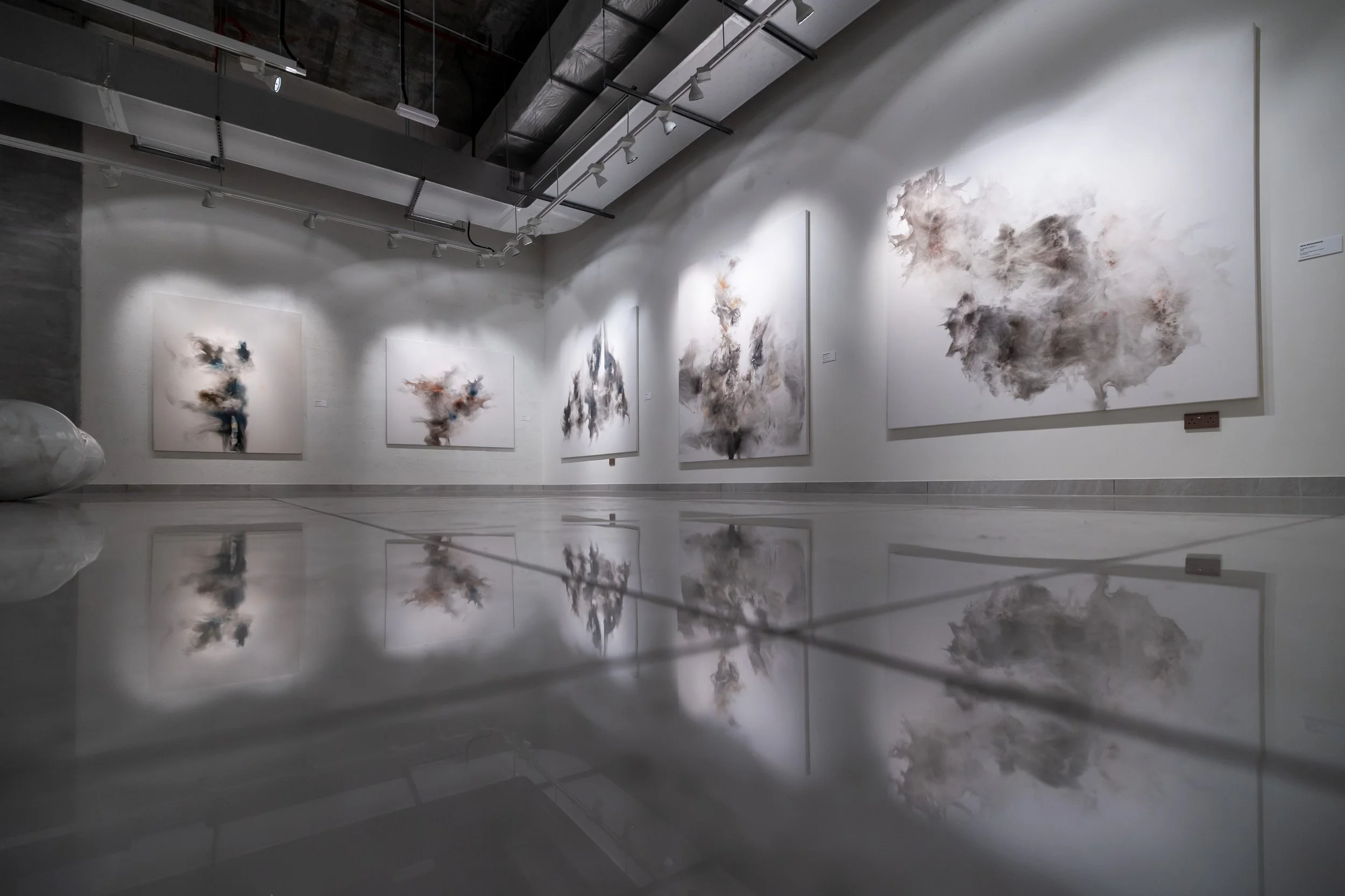 An art gallery with abstract paintings on white walls, featuring dark, smoky, and cloud-like shapes, with a highly reflective floor mirroring the artworks.
