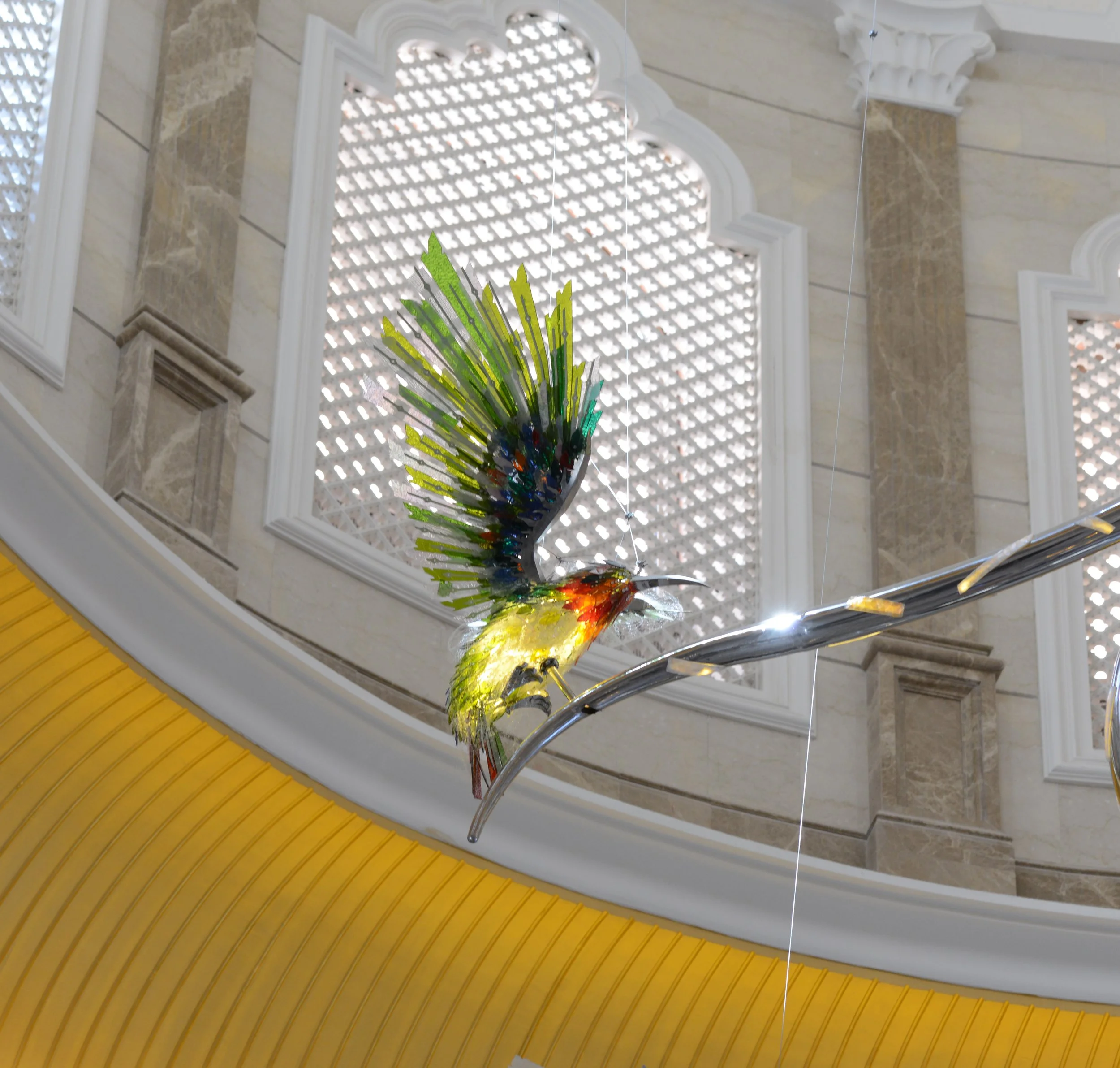 Colorful glass sculpture of a hummingbird with iridescent wings, mounted on a metal branch in an elegant indoor setting with decorative windows.