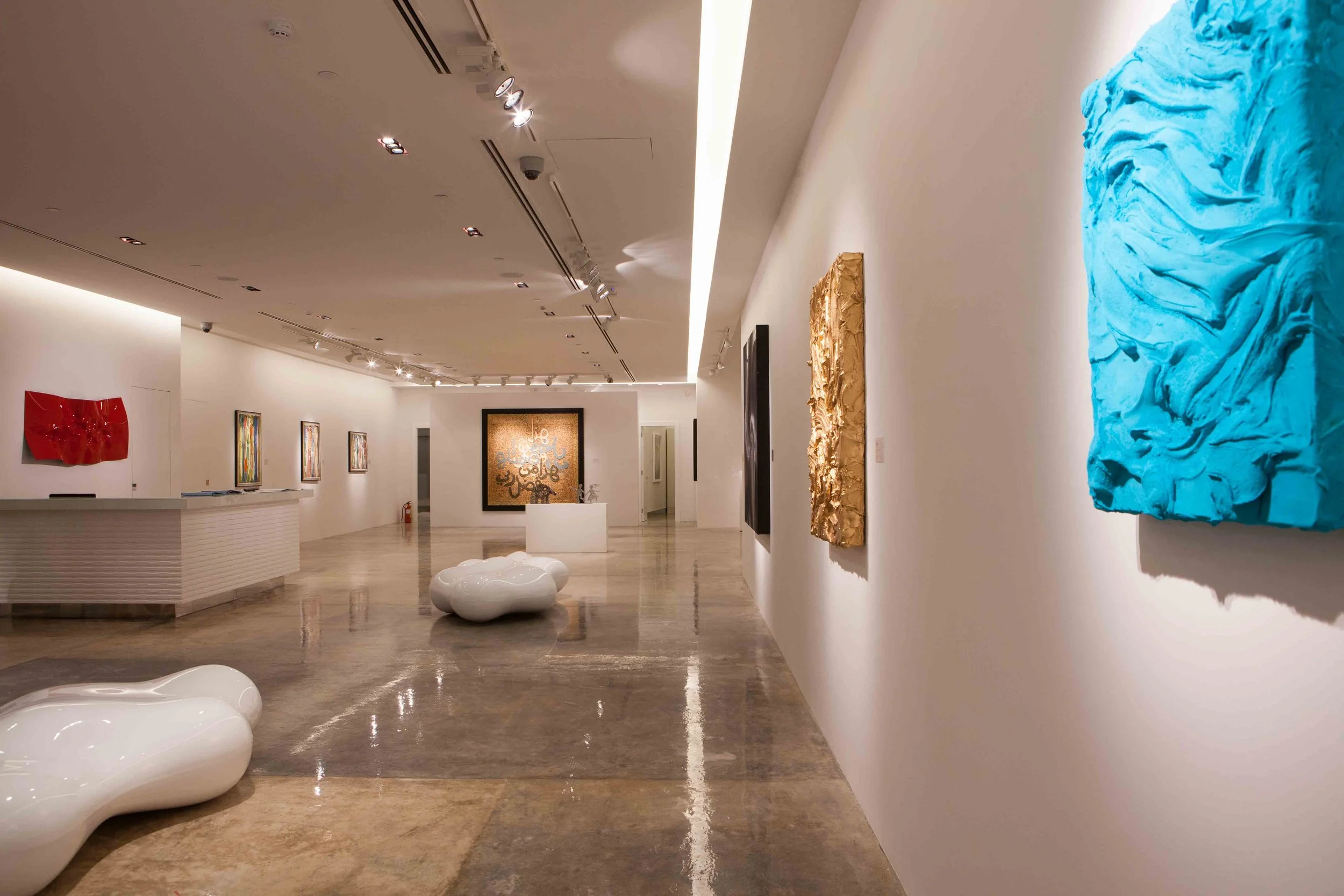 Art gallery with various abstract paintings and sculptures on white walls and polished concrete floor, illuminated by track lighting.