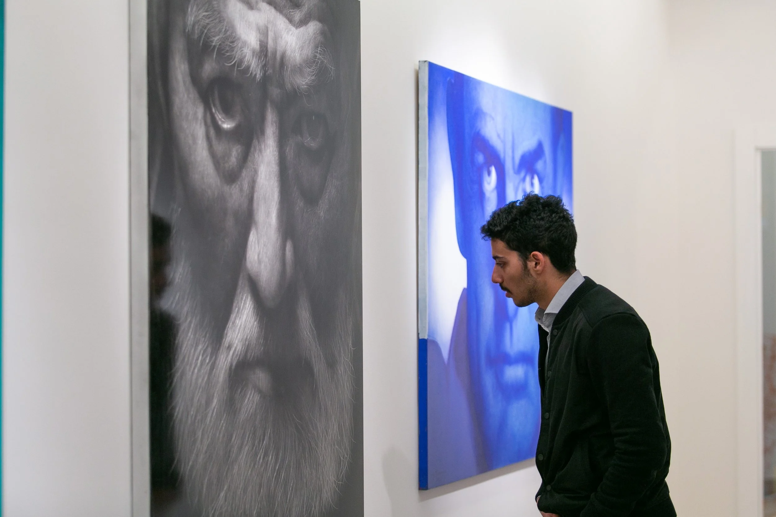 A man with dark, curly hair and a mustache looks at paintings of faces on a white gallery wall, one in black and white and the other in blue.