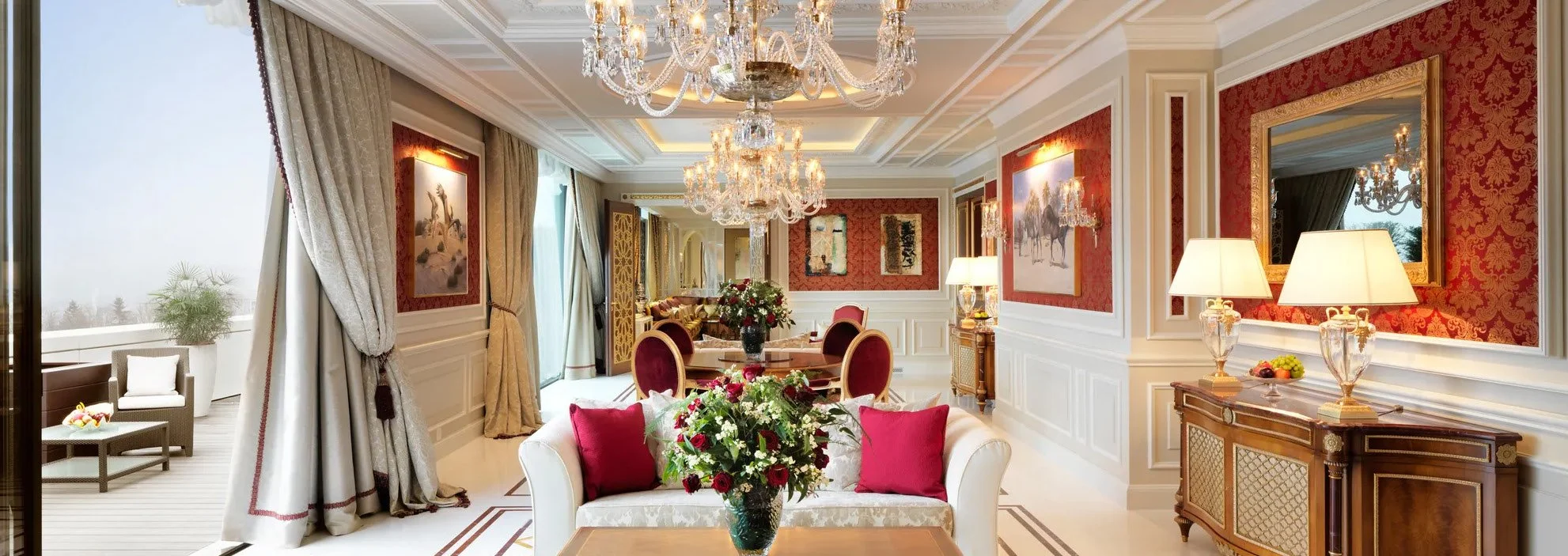 Luxurious living room with elegant furniture, chandeliers, and red and gold decor, leading to an outdoor balcony with patio furniture.