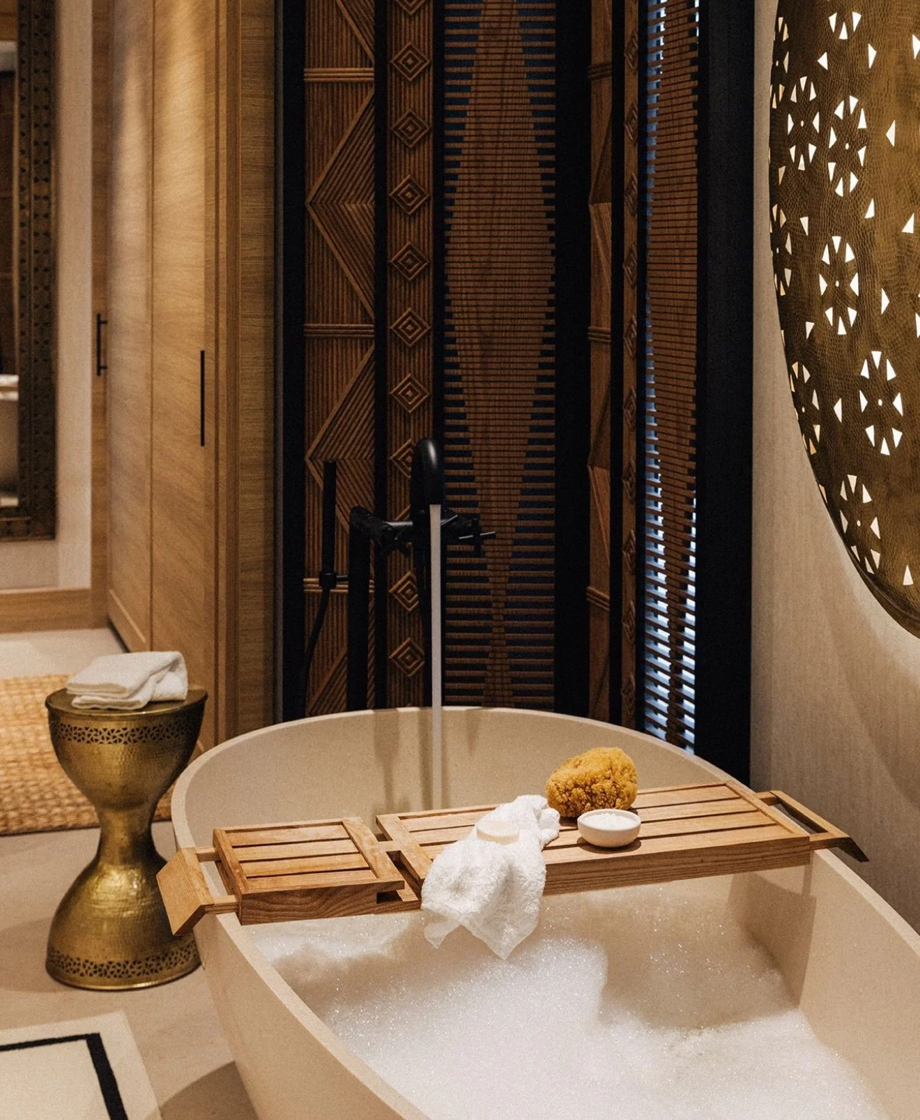 Luxurious bathroom with a freestanding bathtub filled with bubbles, a wooden bath tray holding a towel and a yellow bath sponge, and decorative wooden walls with intricate patterns and window blinds.