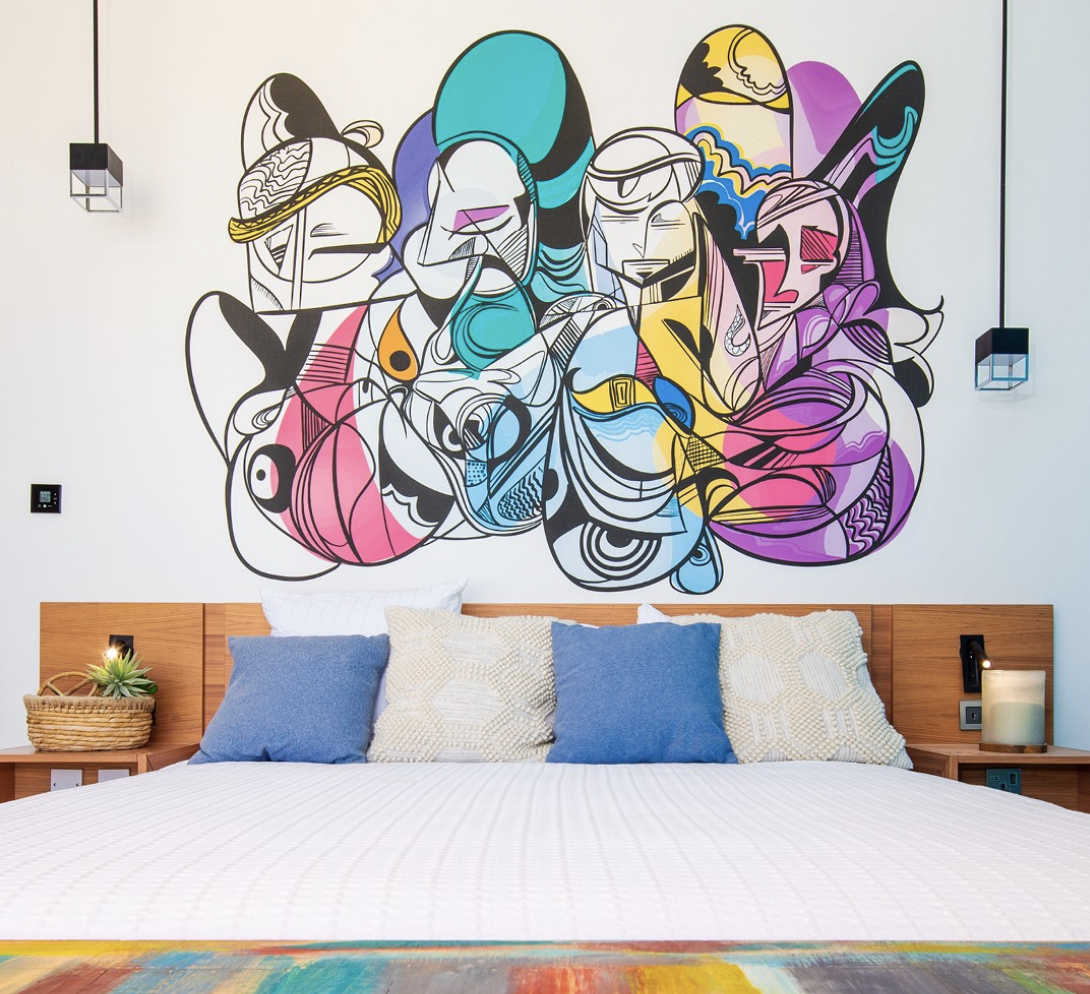 Modern bedroom with a colorful abstract mural of three women above the bed, which has four pillows and a wooden headboard, decorated with plants and a candle.