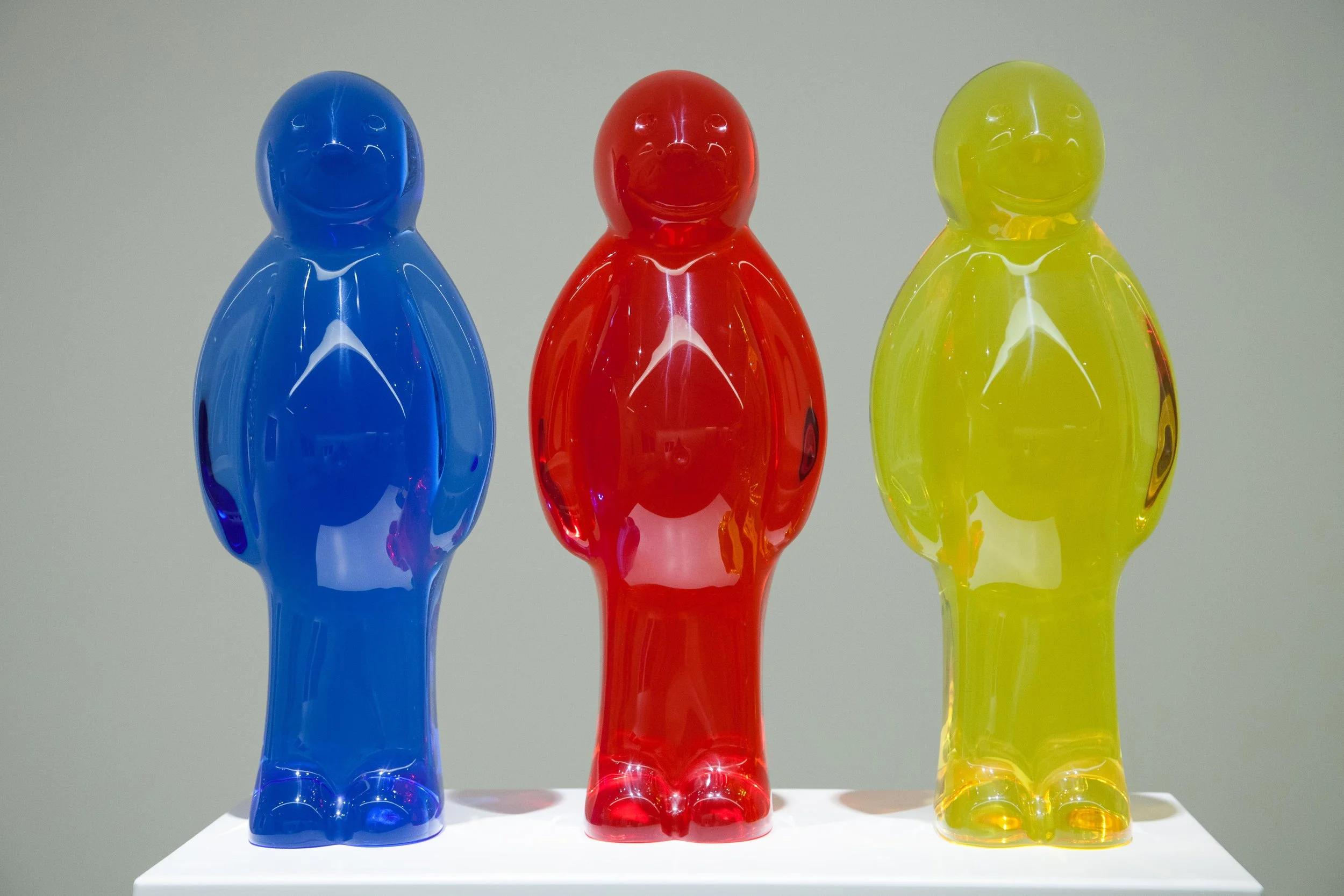 Three colorful glass figurines shaped like people, in blue, red, and yellow, standing on a white display table against a plain background.