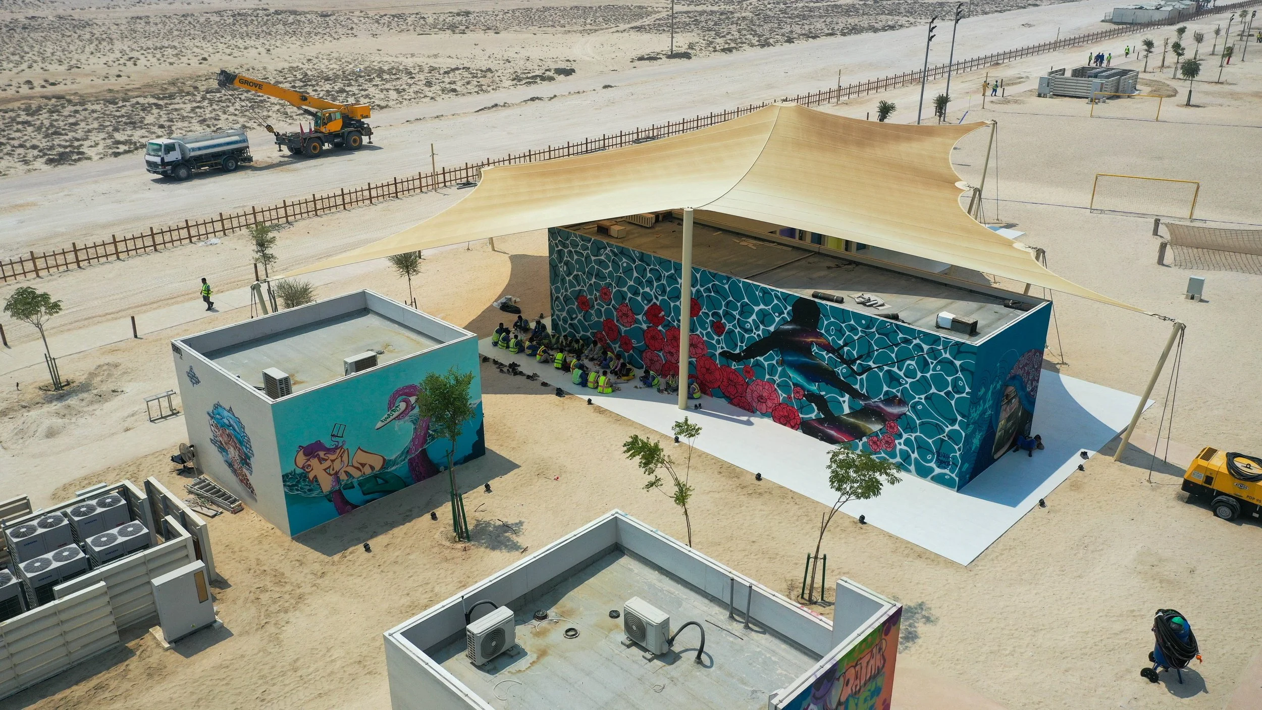 Aerial view of a modern outdoor skatepark with painted murals on the walls, covered by a large fabric canopy, and surrounded by small trees, construction equipment, and sandy ground.