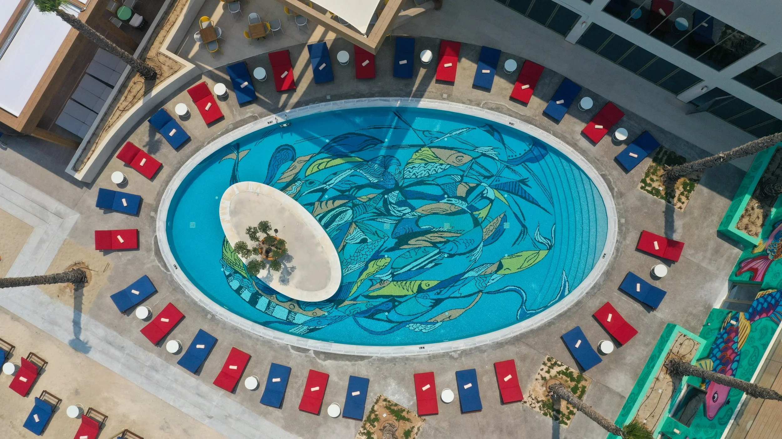 A swimming pool with a colorful abstract design on the bottom, surrounded by red and blue lounge chairs and small white tables, in an outdoor courtyard area.