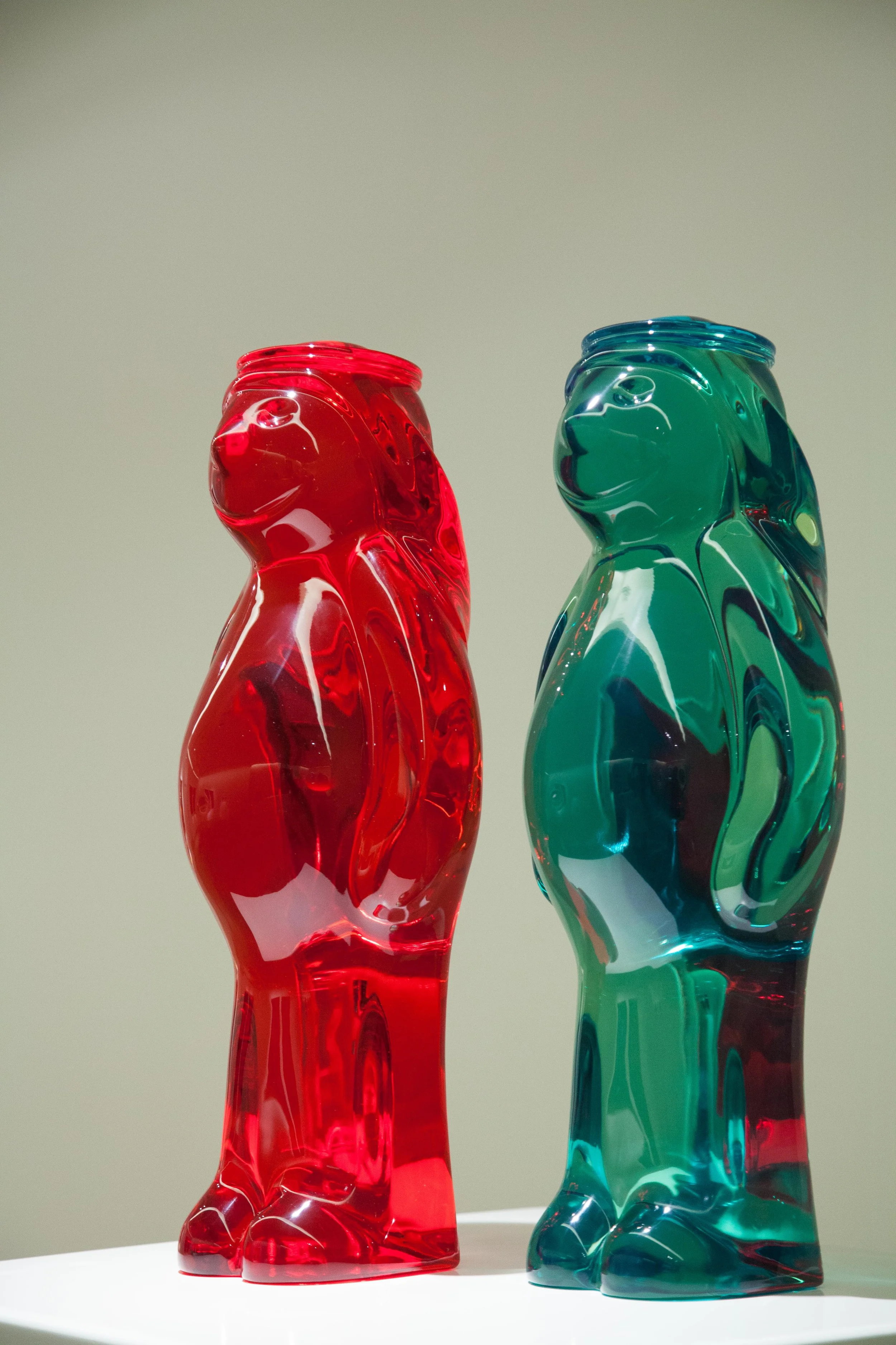 Two glass cat sculptures, one red and one green, positioned side by side against a plain background.