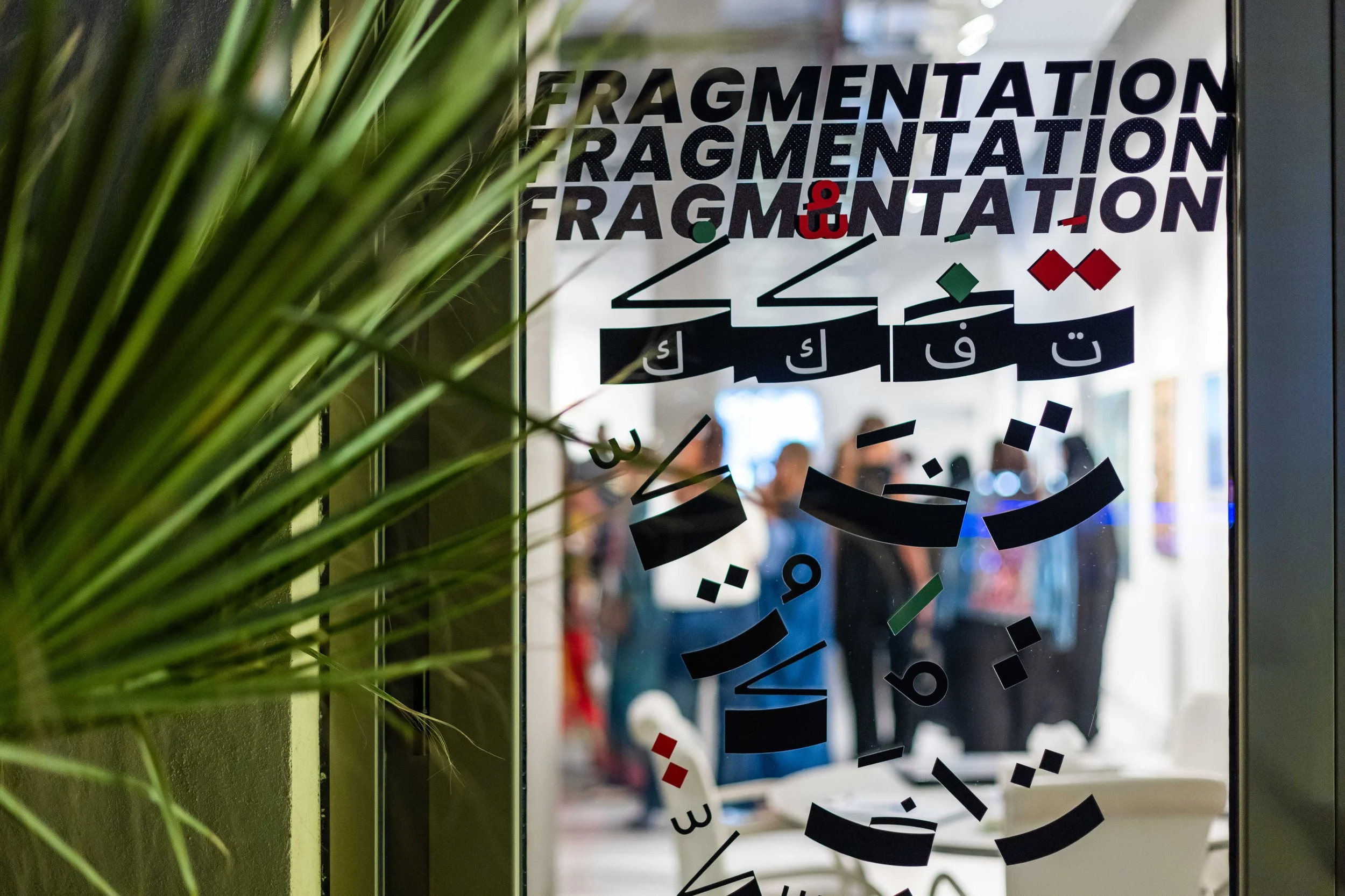 View through a glass door with decorative paper cutouts in black, red, green, and white in the shape of Arabic letters and numerals, with the word "FRAGMENTATION" repeated at the top. People are visible in the background, blurred, standing inside an 