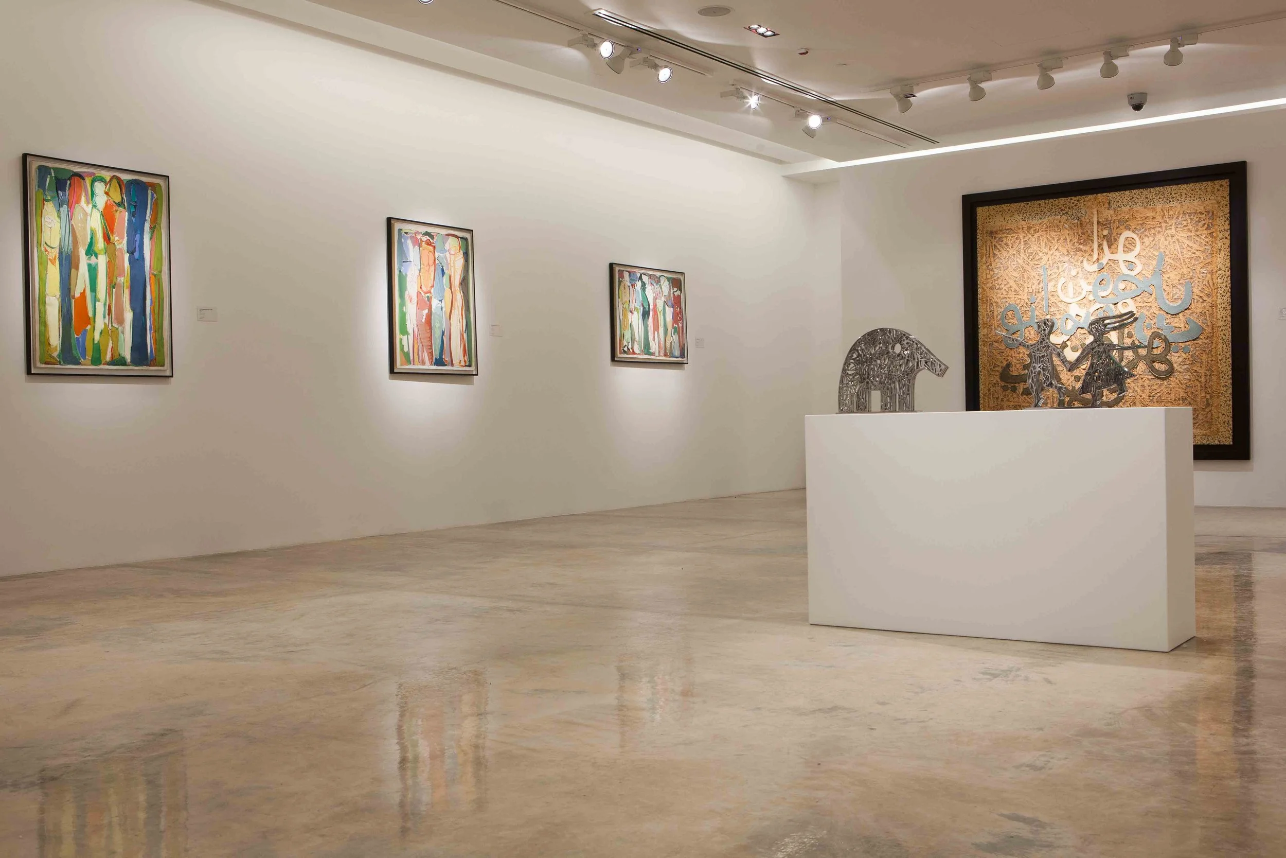 An art gallery with four colorful abstract paintings on the left wall, two metal sculptures on the white pedestal in the center, and a large framed artwork with Arabic calligraphy in the background. The gallery has a polished marble floor and track l