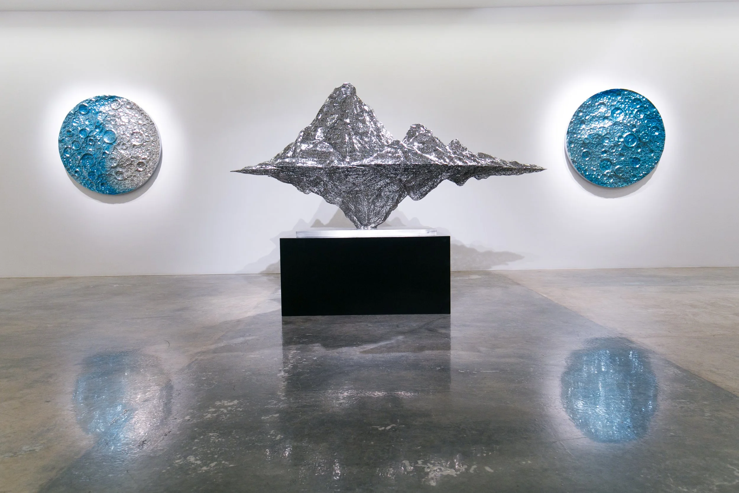 Contemporary art gallery with a metallic mountain sculpture on a black pedestal in the center and two circular wall-mounted artworks with blue and silver textures on either side.